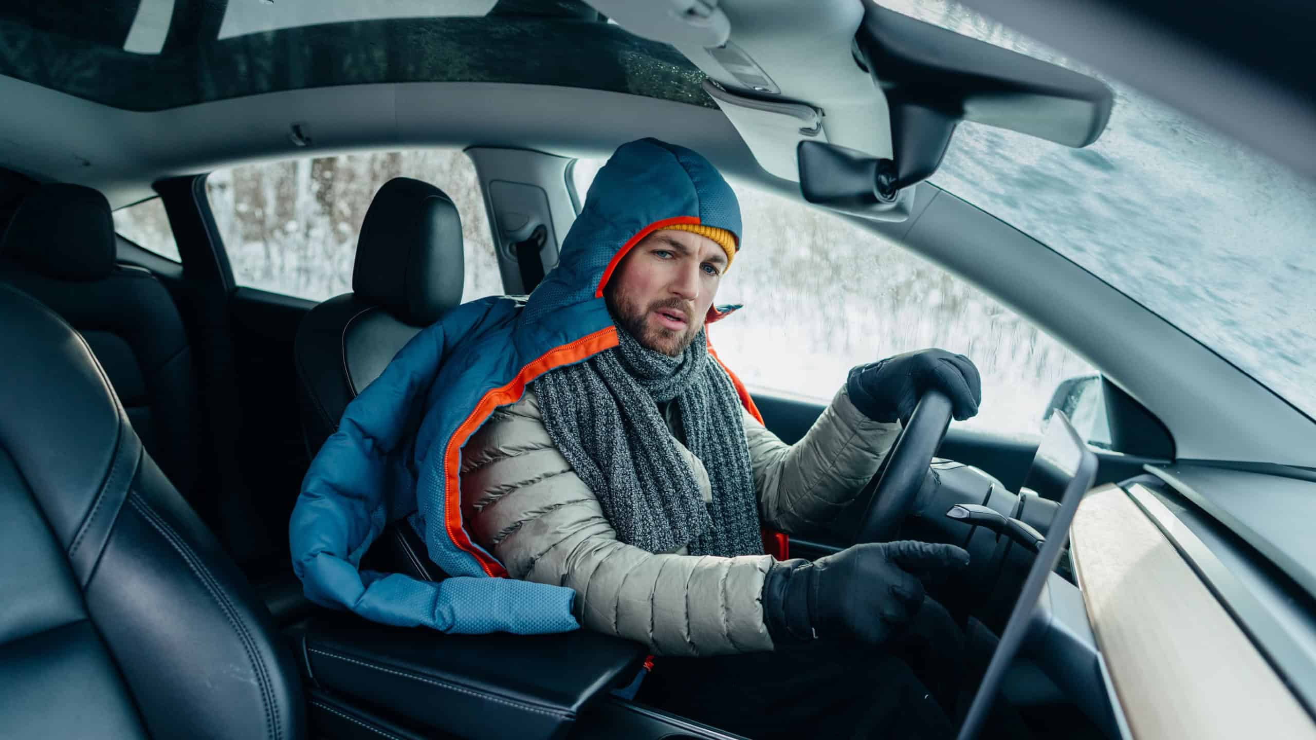 Driver experiencing extreme cold inside an electric car, wearing a sleeping bag and winter clothes to conserve energy