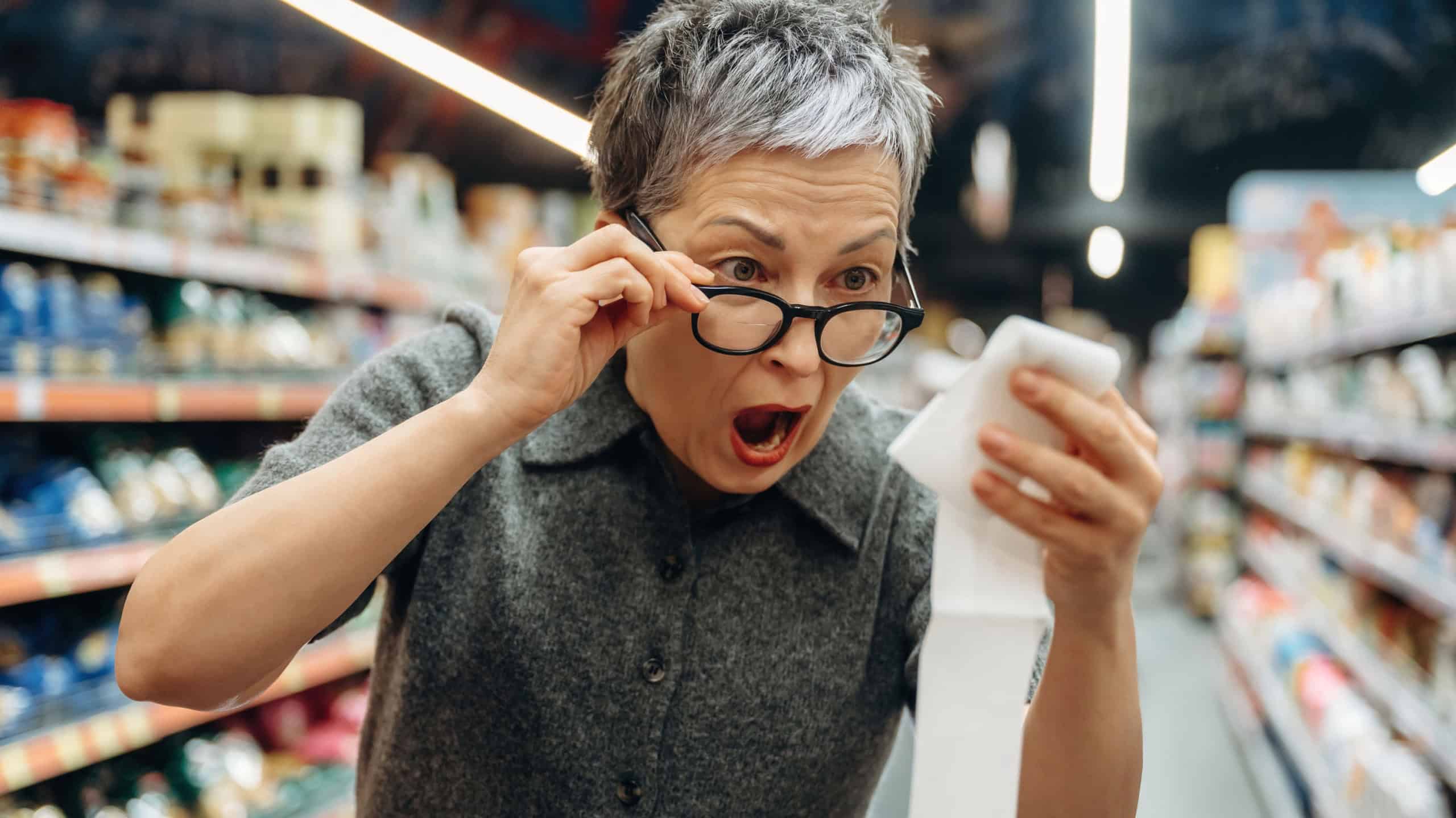 Mature woman with gray hair and glasses reacts in surprise while checking a lengthy receipt in a grocery store aisle with various products around her