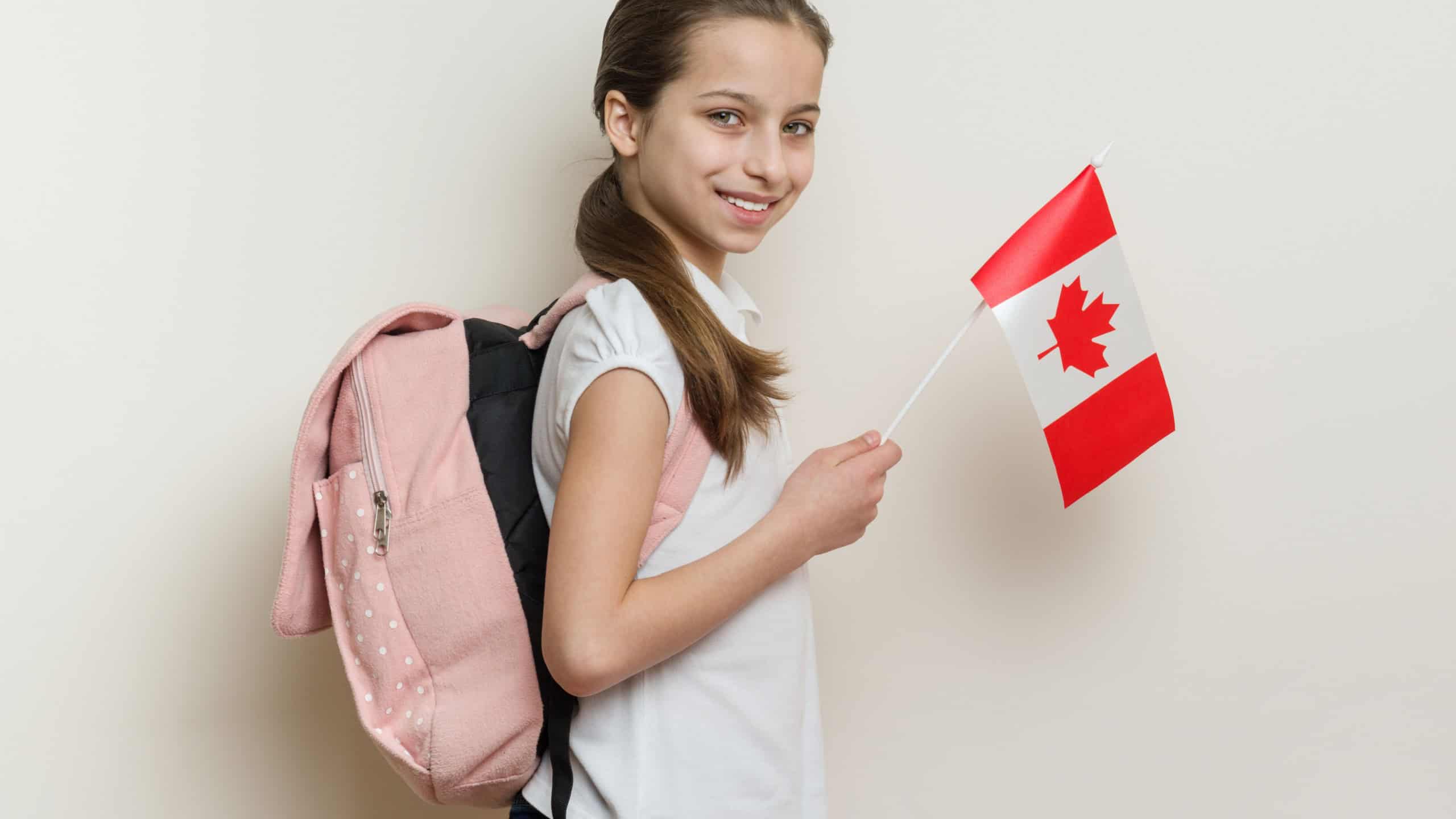 Schoolgirl 10 years in white T-shirt with a backpack holding the flag of Canada, background bright wall in the school.