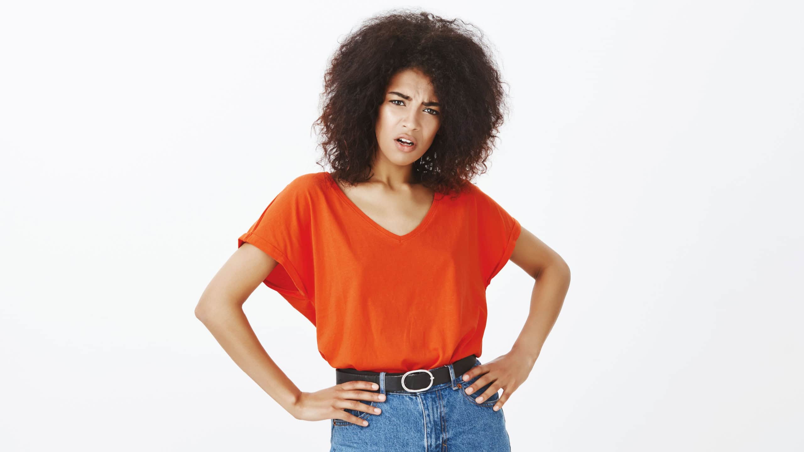 Indoor shot of offended attractive woman with curly hair holding hands on hips and staring with displeased insulted expression at camera, being shocked with offencive and rude words