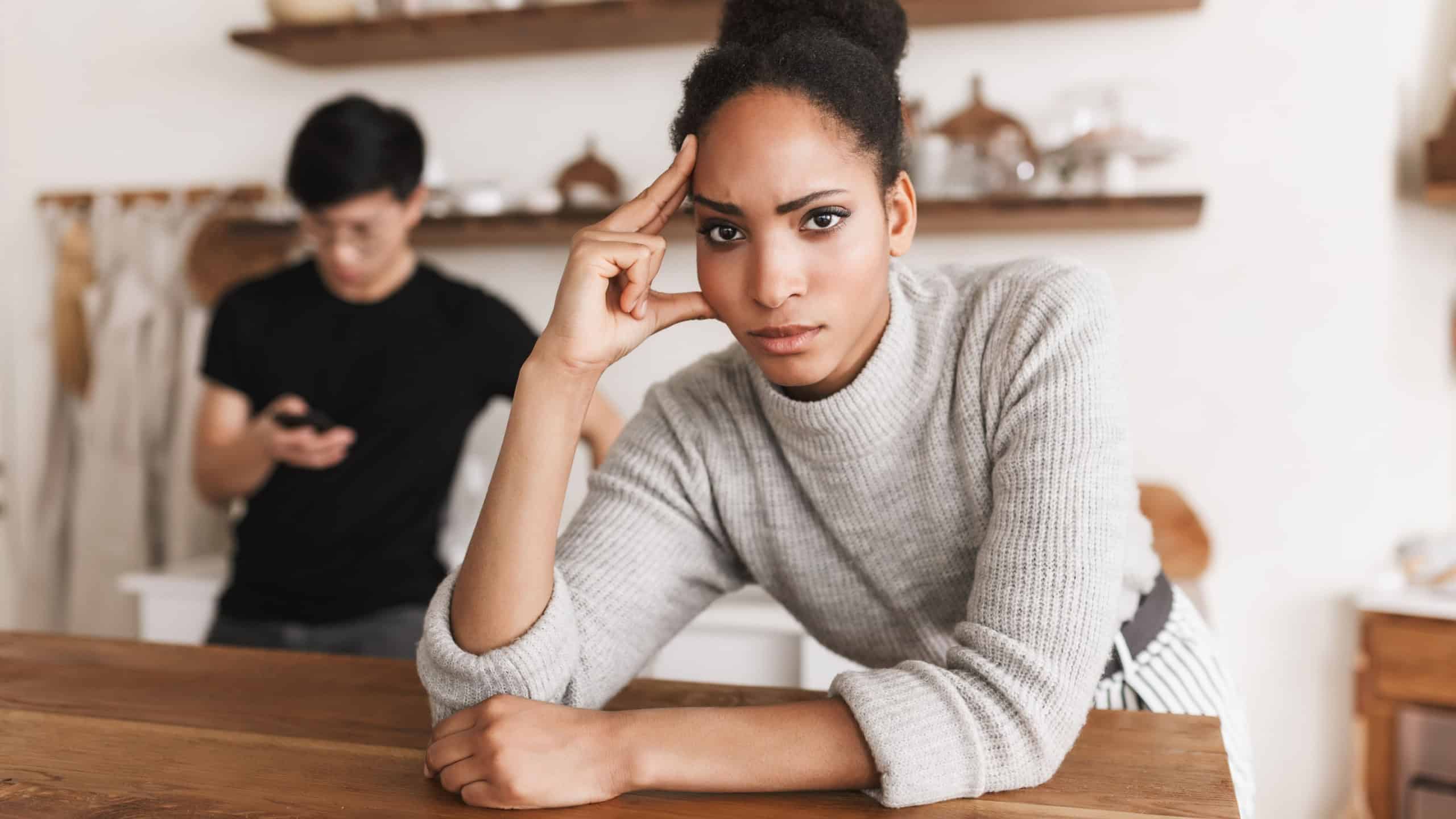 Serious african american woman holding hand near head angrily looking in camera with asian man using cellphonr on background. Young international couple in quarrel spending time on kitchen at home