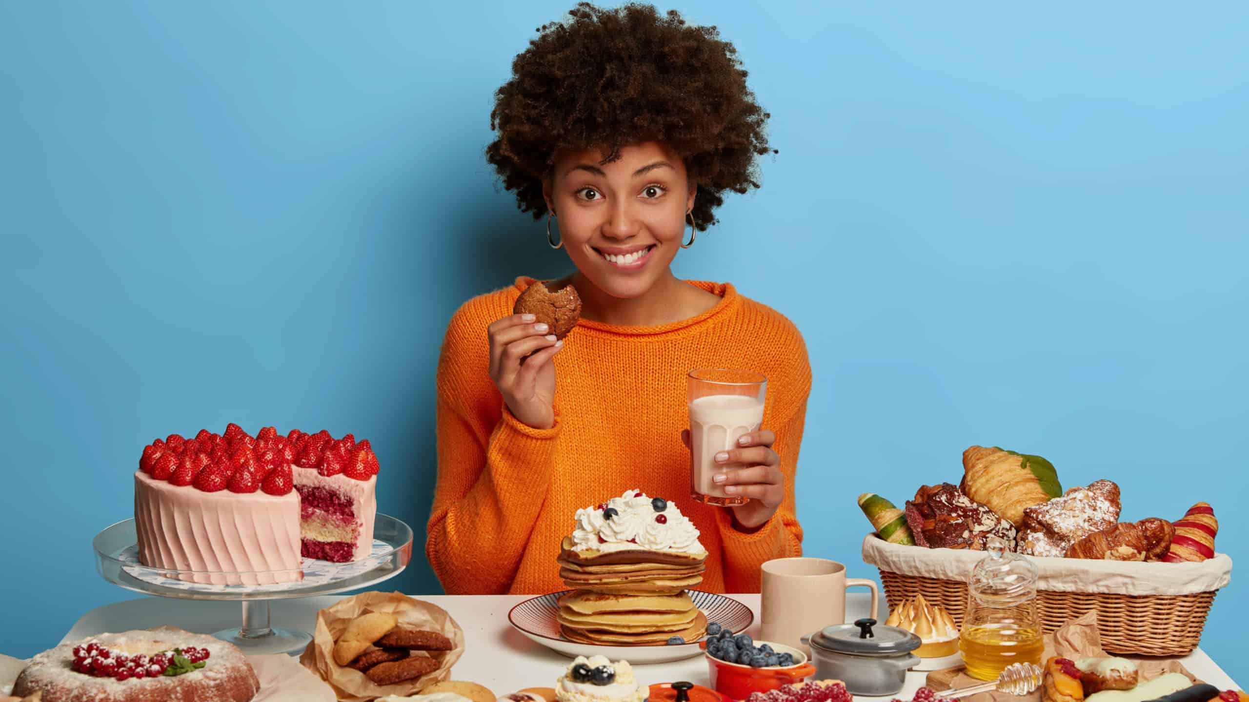 Glad smiling woman with Afro curly hairdo eats tasty pastry with milk, has good mood to eat delicious desserts, tries yummy just baked cookies, cannot loose weight because of sugar addiction