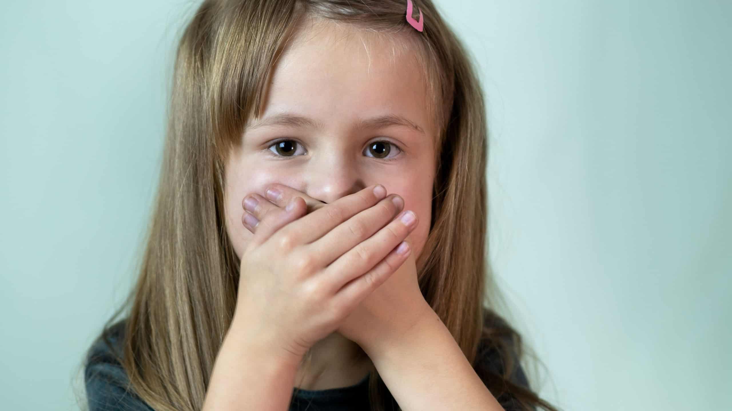 Close-up portrait of little child girl with long hair covering her mouth with hands.