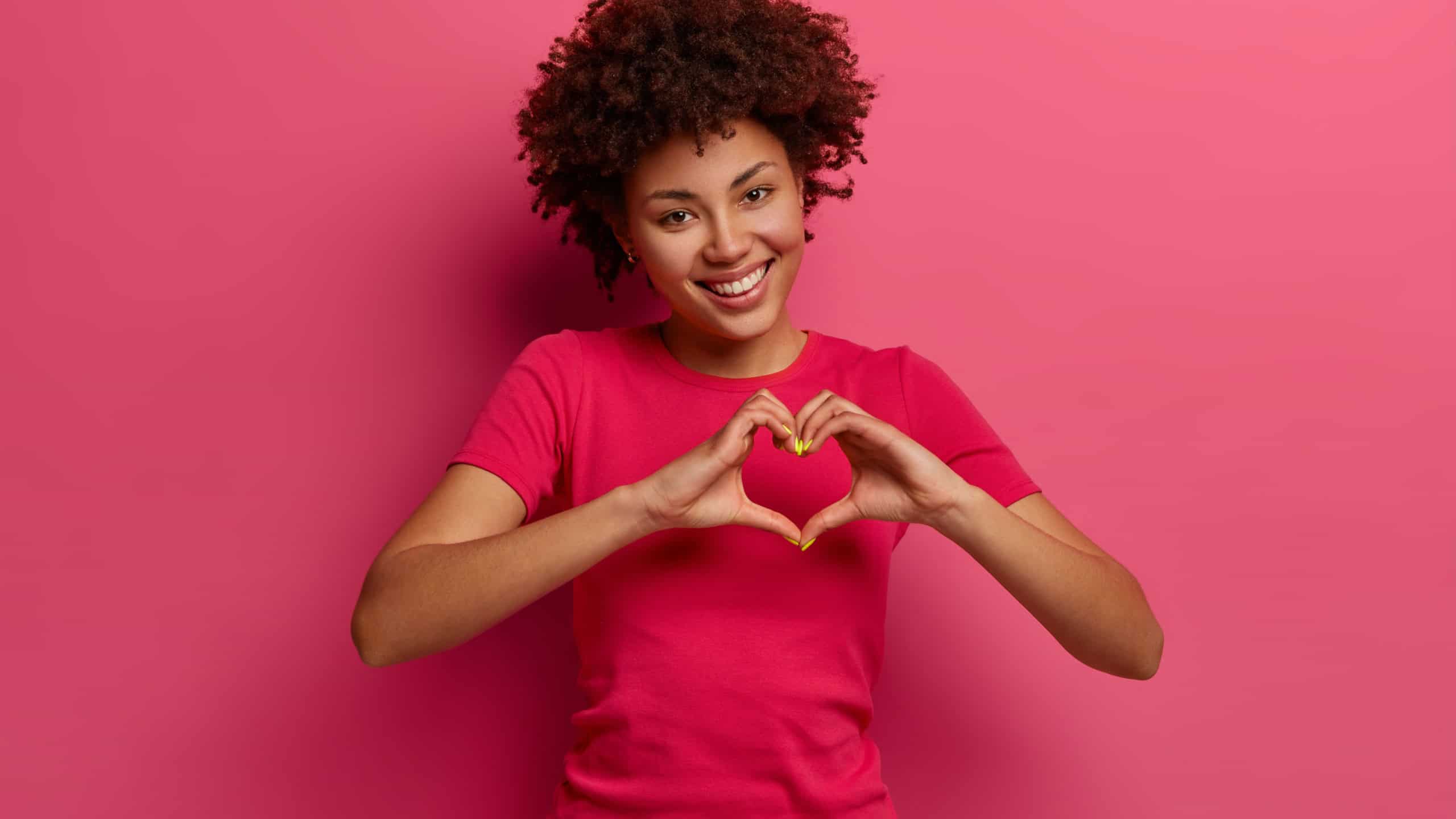 Pretty curly African American woman confesses in love, makes heart gesture, shows her true feelings, has happy expression, wears casual red t shirt, poses over pink background. Relationship concept