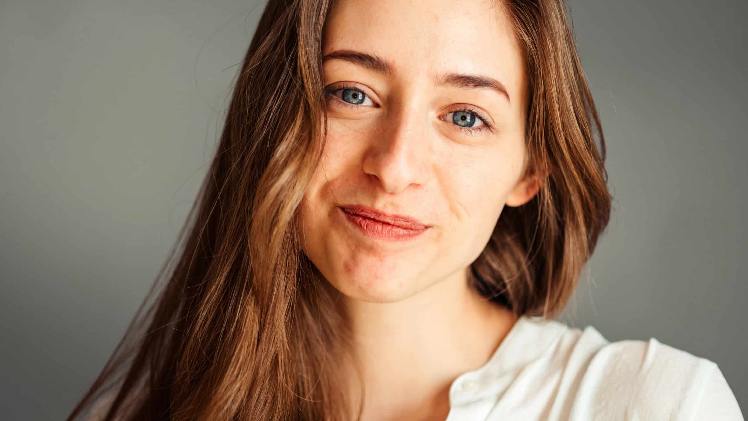 Close-up portrait of a smiling young girl in a white shirt on a gray background. Hands near the face. without retouching and makeup.