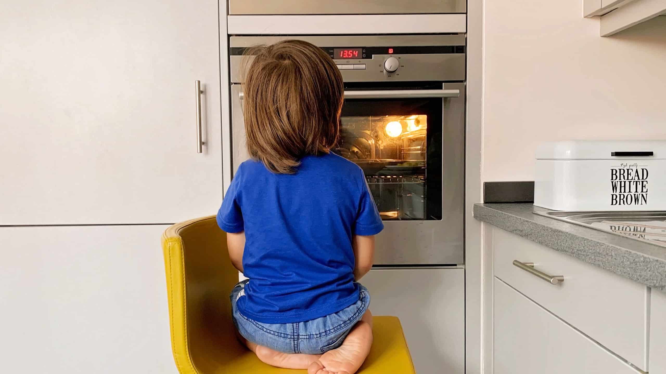 Little hungry toddler boy sitting at oven looking inside through the glass waiting for pizza.