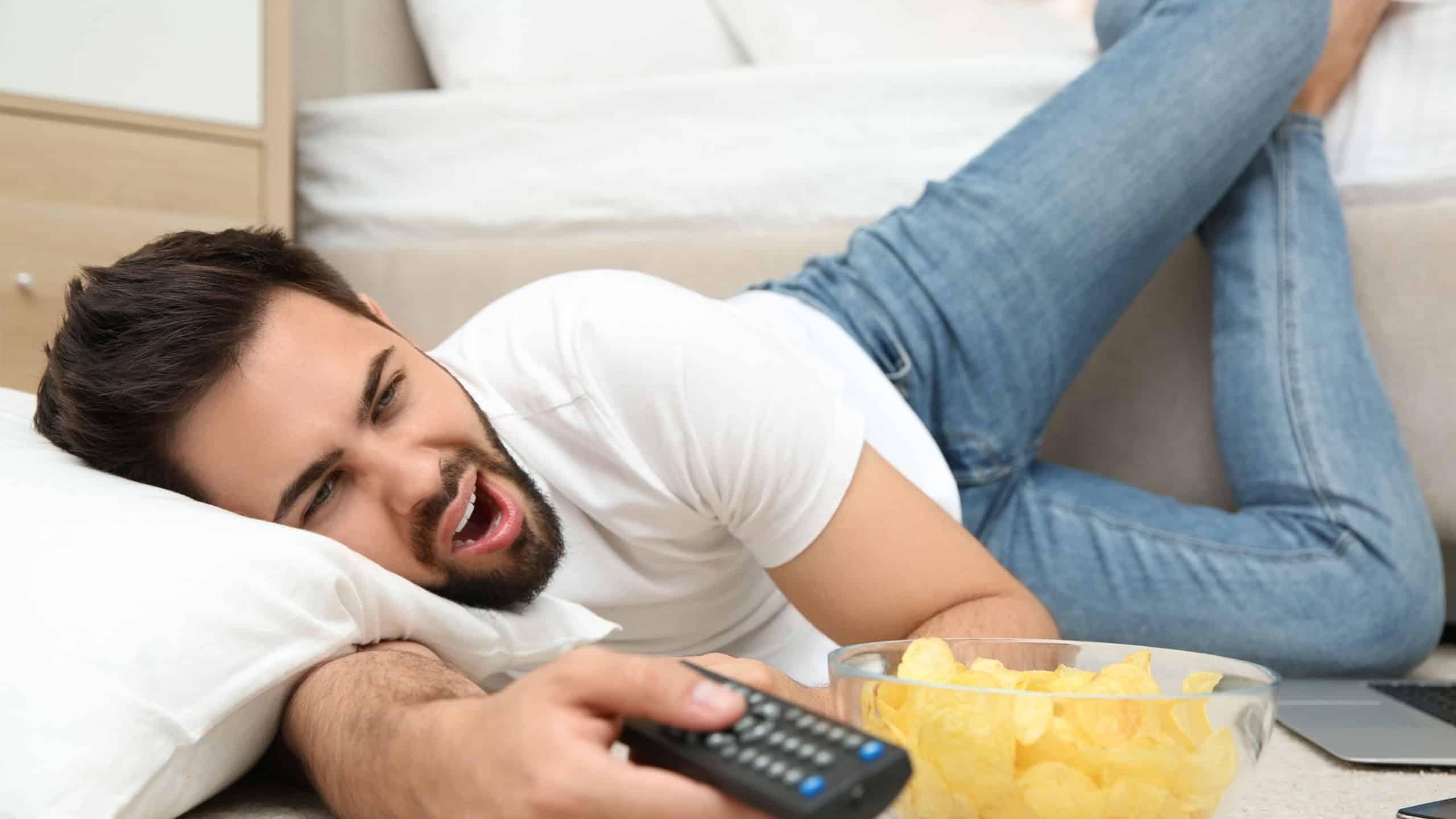Lazy young man with bowl of chips watching TV while lying on floor at home