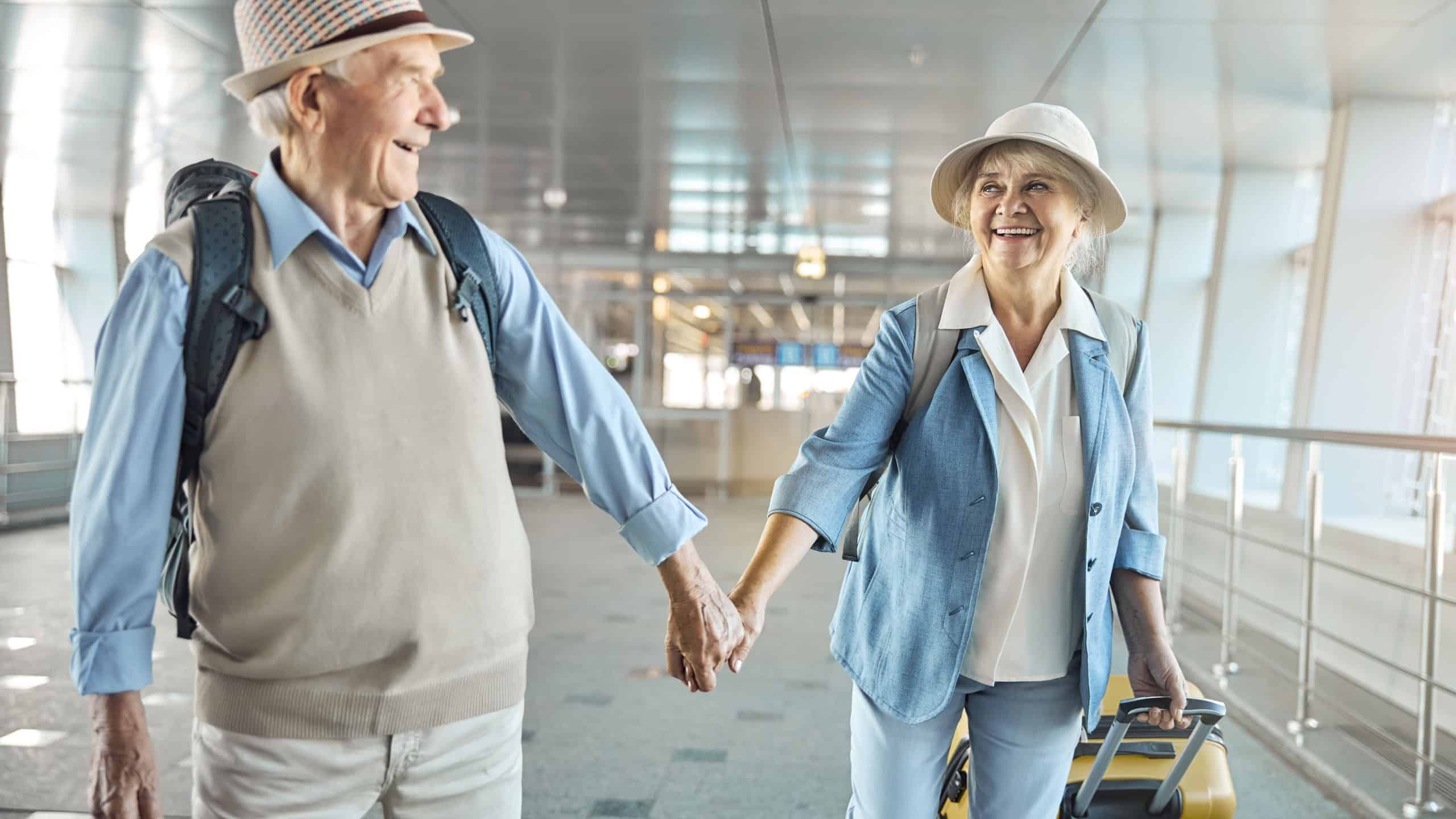 Pleased gray-haired male tourist smiling at a joyful attractive elderly lady with a trolley suitcase