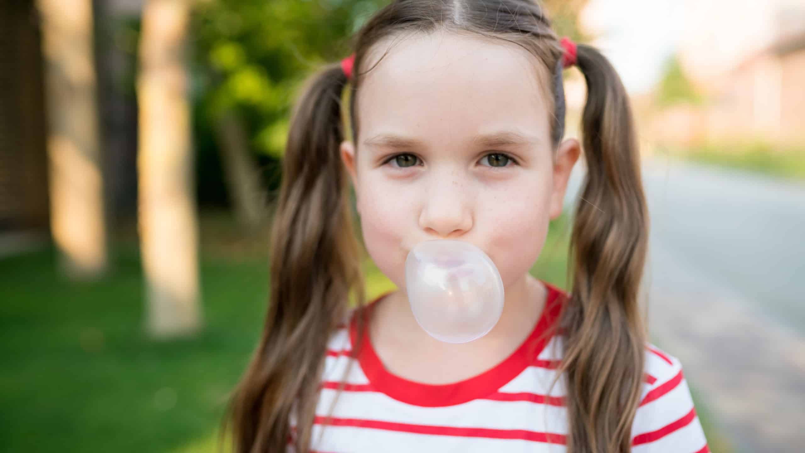 Cute child girl with two ponytails looking at camera and inflating bubble with chewing gum outdoor. Kids blowing bubble gum.