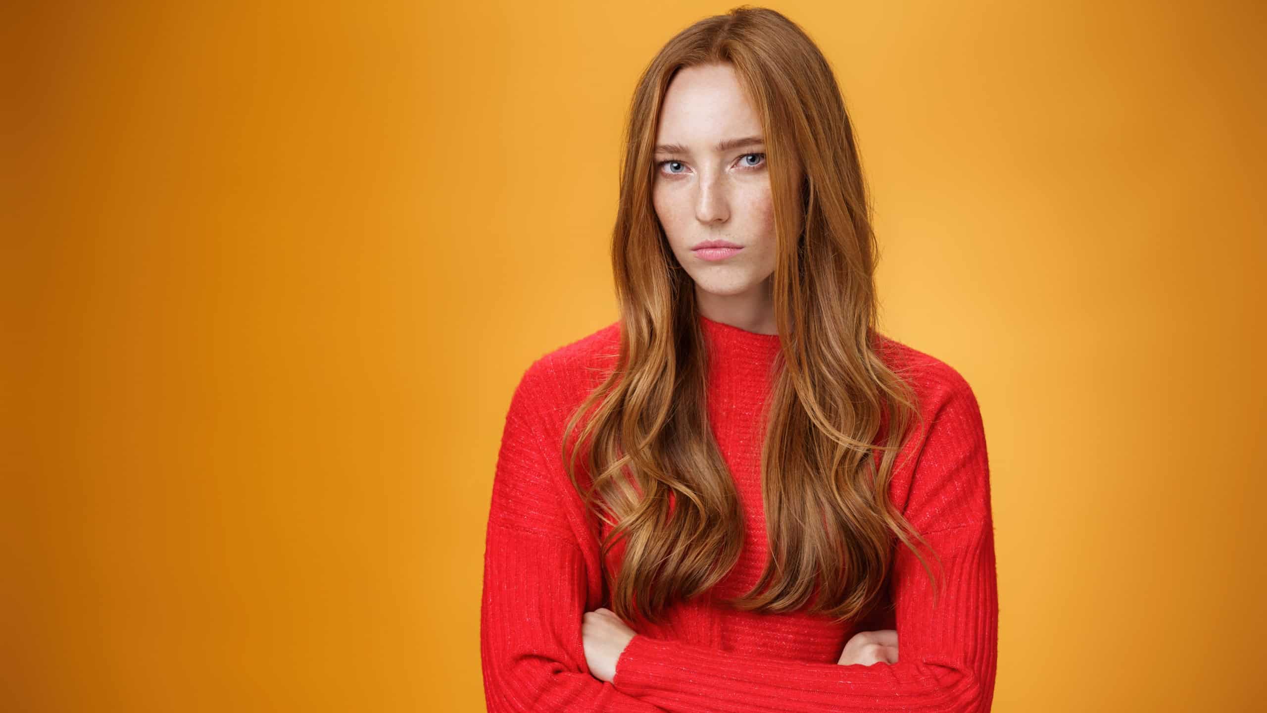 Suspicious intense and defensive ginger girl standing in passive-aggressive pose pouting and frowning looking with disbelief and disdain at camera, offended against orange background