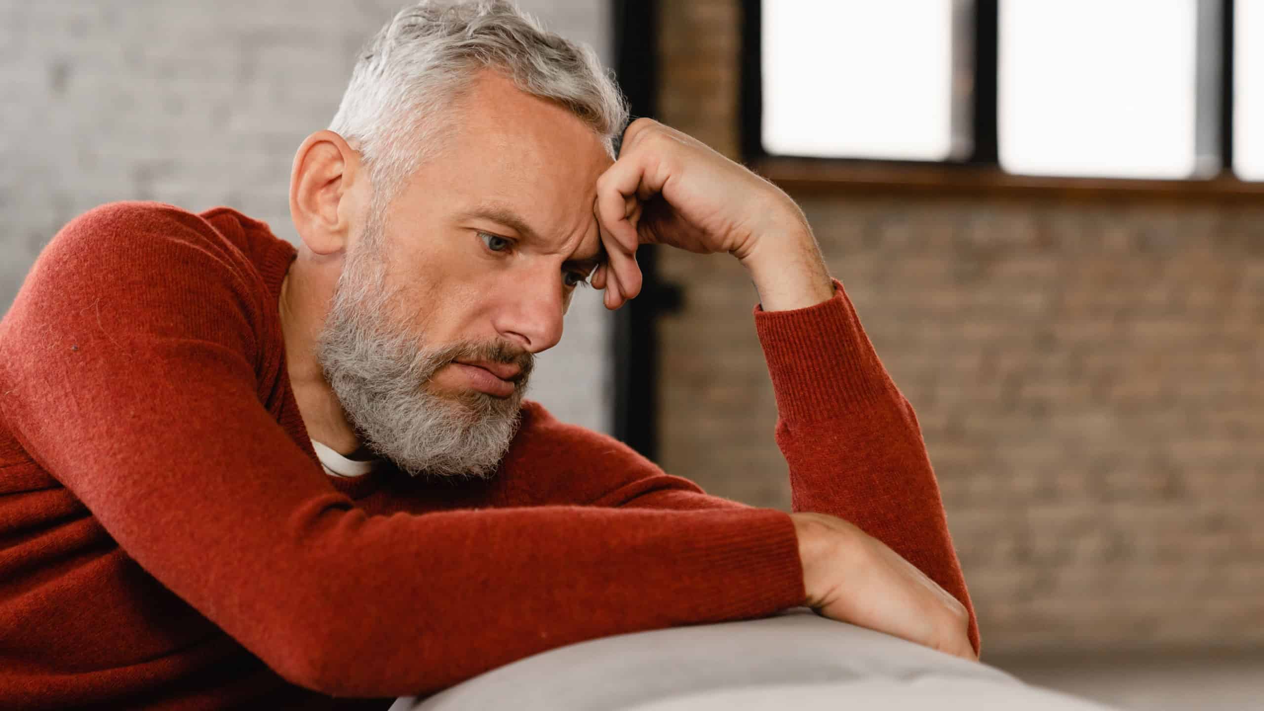 Sad stressed troubled mature middle-aged man sitting on the couch, missing his friends, feeling bad unwell unhealthy, suffering from depression at home