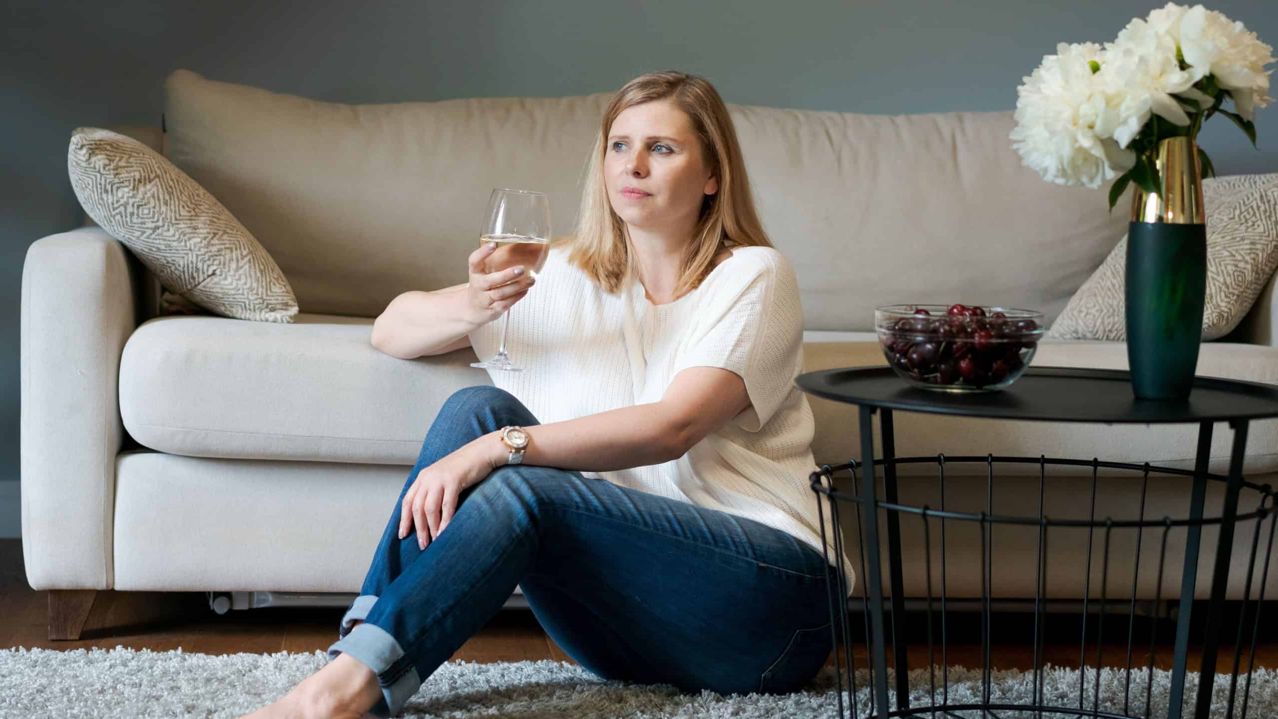 Fashionable woman. Blond caucasian woman holding a glass of wine looking towards the window while sitting on the carpet on the floor near the sofa in the living room. Pensive lady resting at home