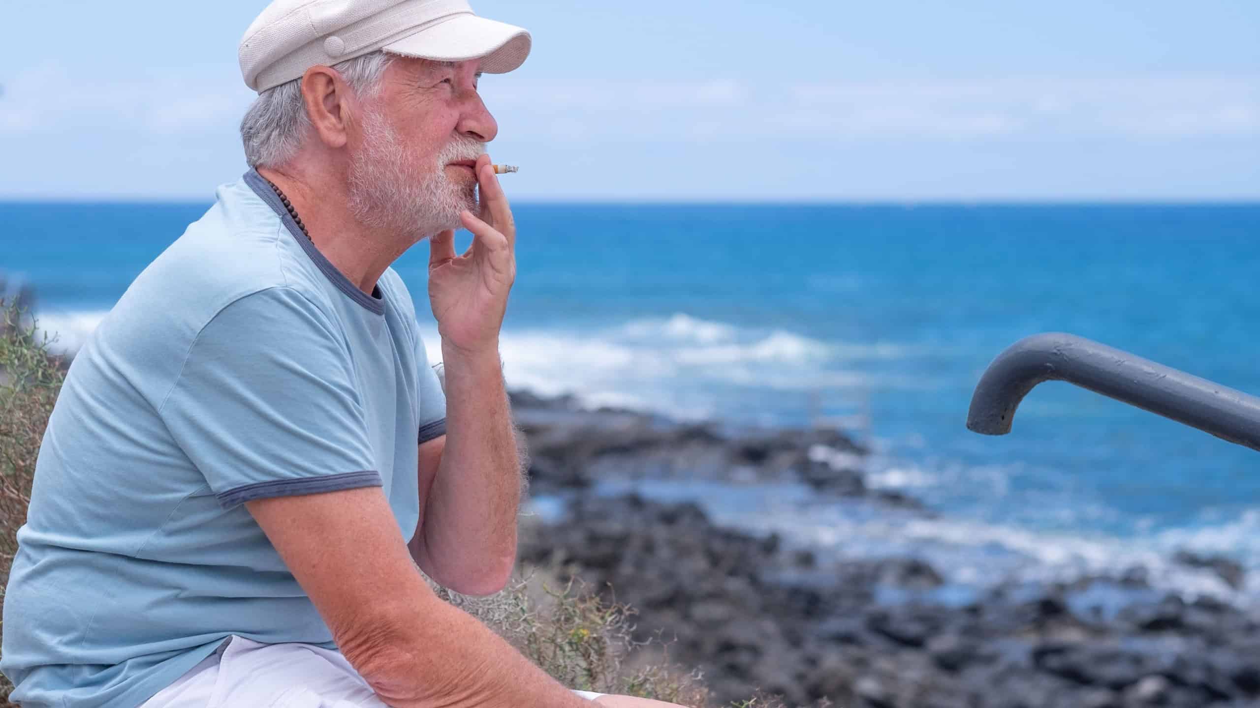Caucasian bearded senior man with hat sitting at the beach looking at the horizon while smoking a cigarette. Horizon over the water, copy space