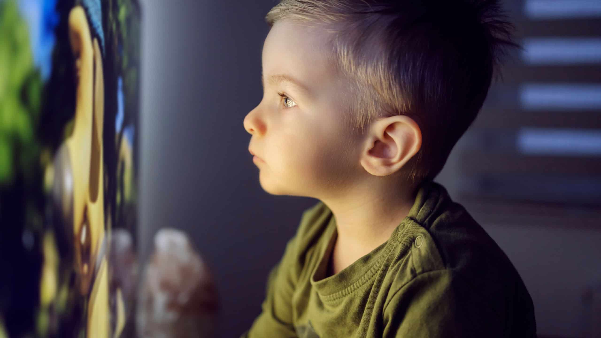 . Close up shot of a baby boy is sitting right in front of the TV and staring at a cartoon screen.