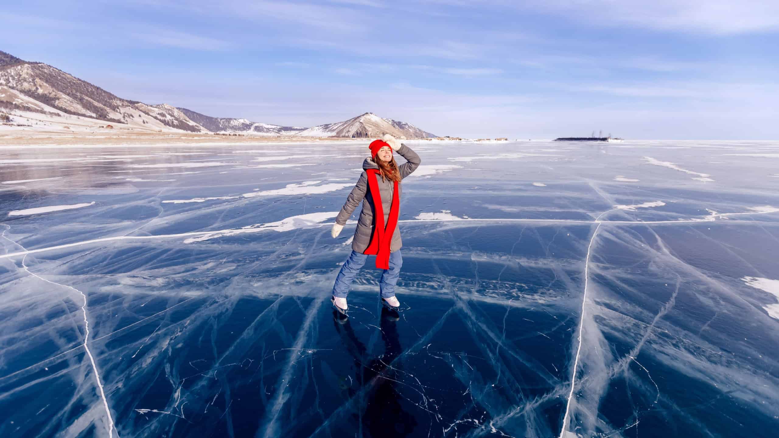 Cheerful woman tourist skates in red hat and scarf on frozen ice of Lake Baikal, sunny winter day.
