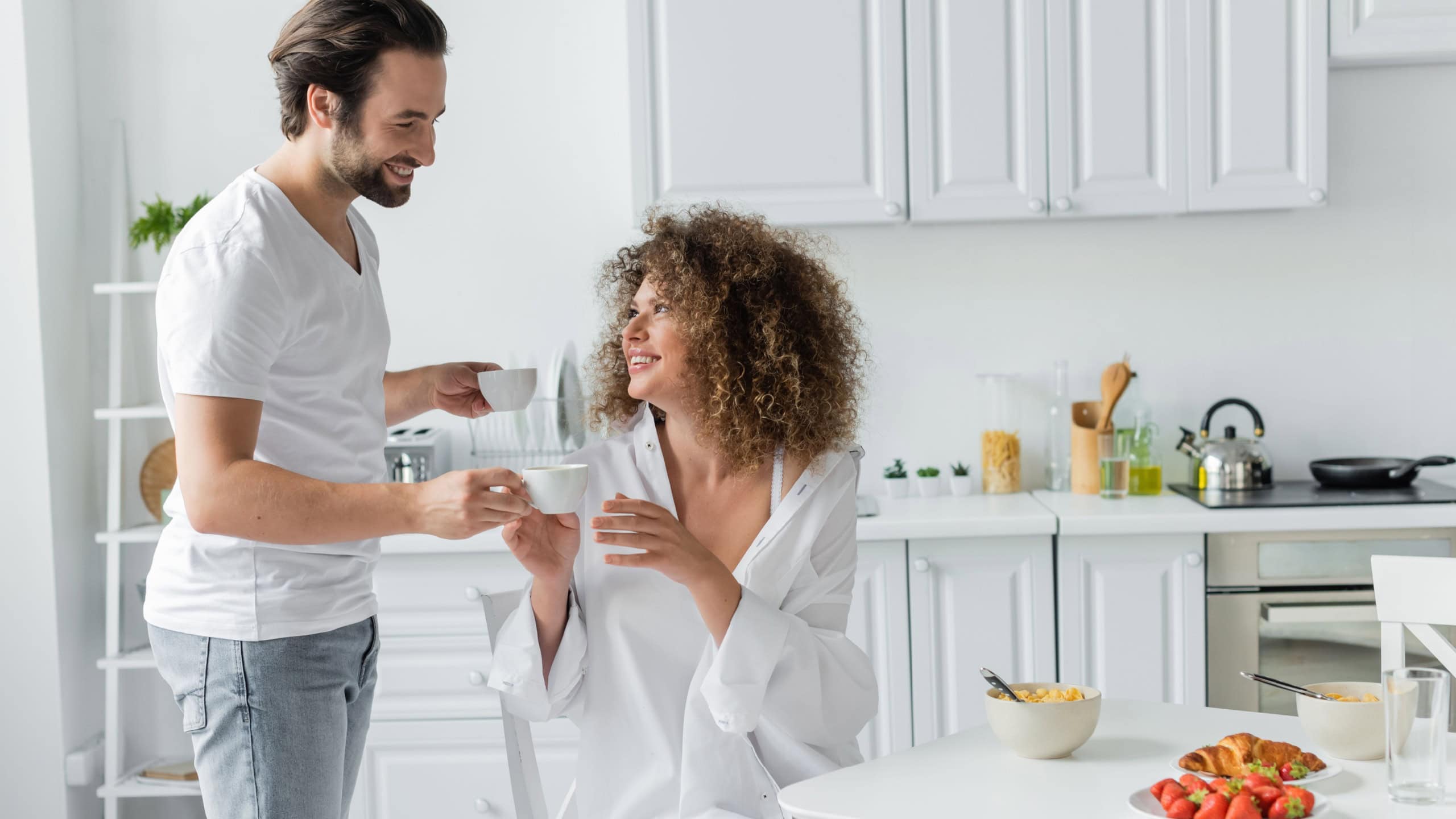 happy young man giving cup of coffee to curly girlfriend during breakfast in kitchen