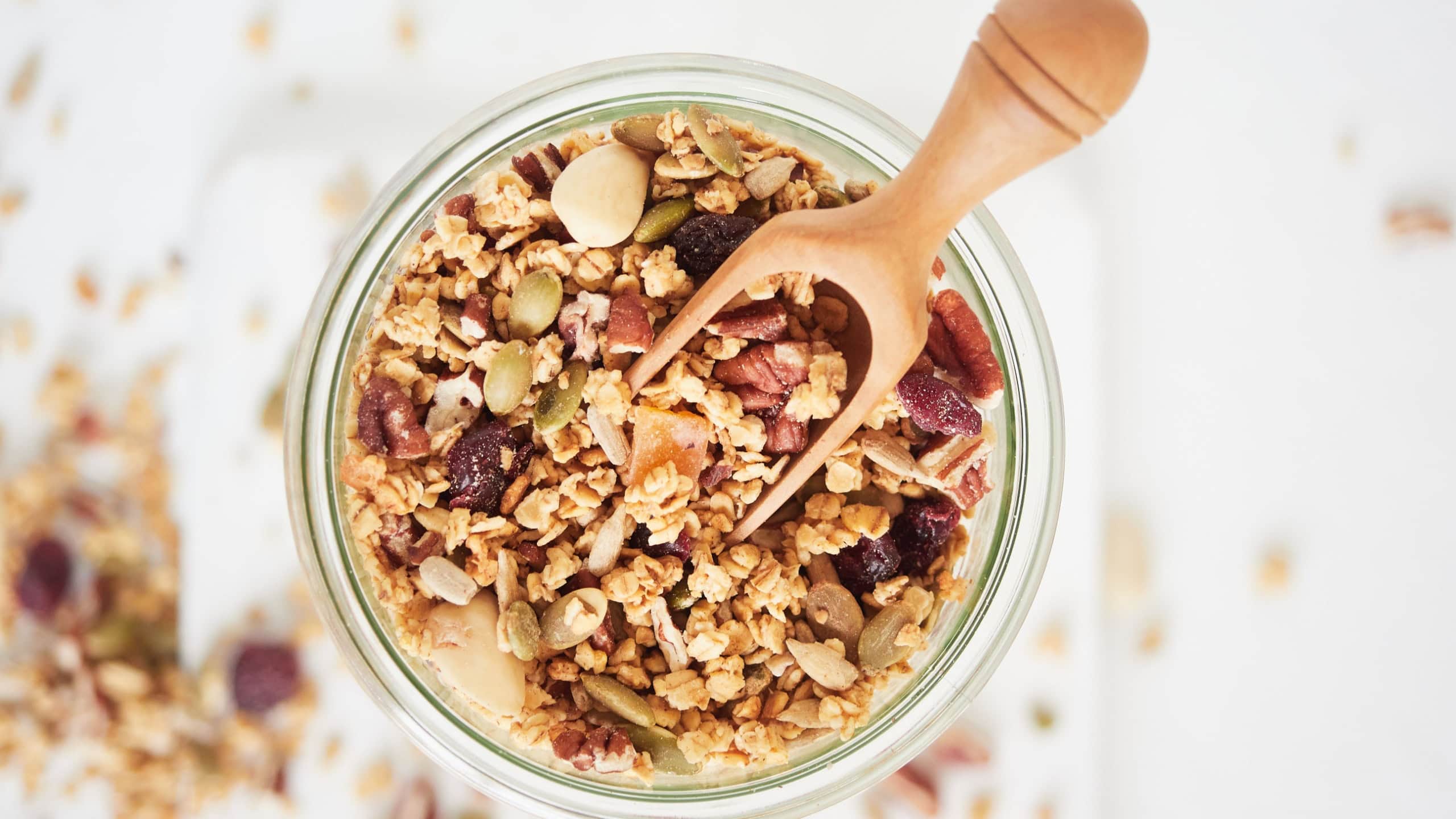 Top view of glass jar with delicious granola with nuts and berries near wooden spoon placed on white table