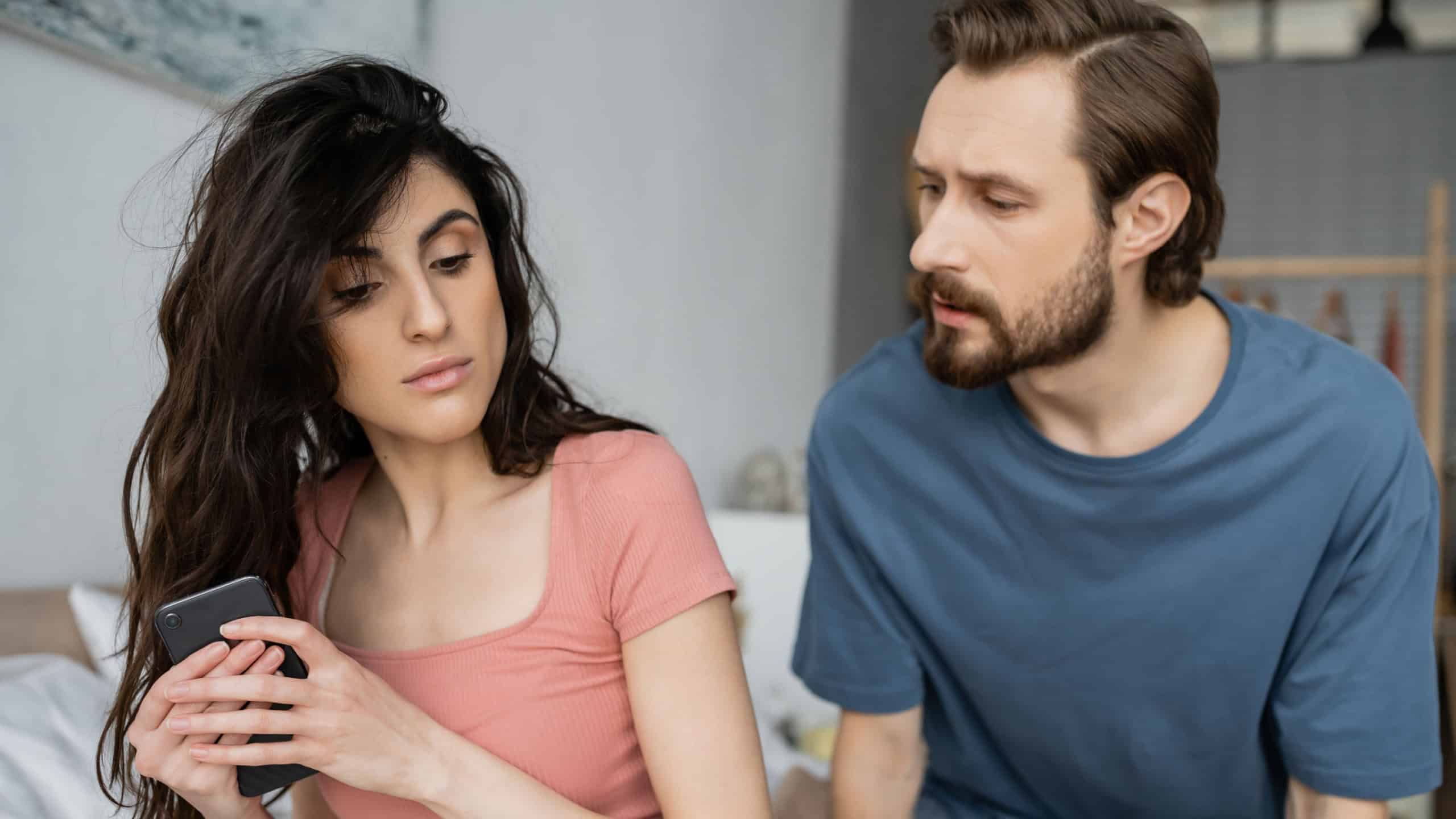 Brunette woman hiding cellphone near jealous boyfriend in bedroom