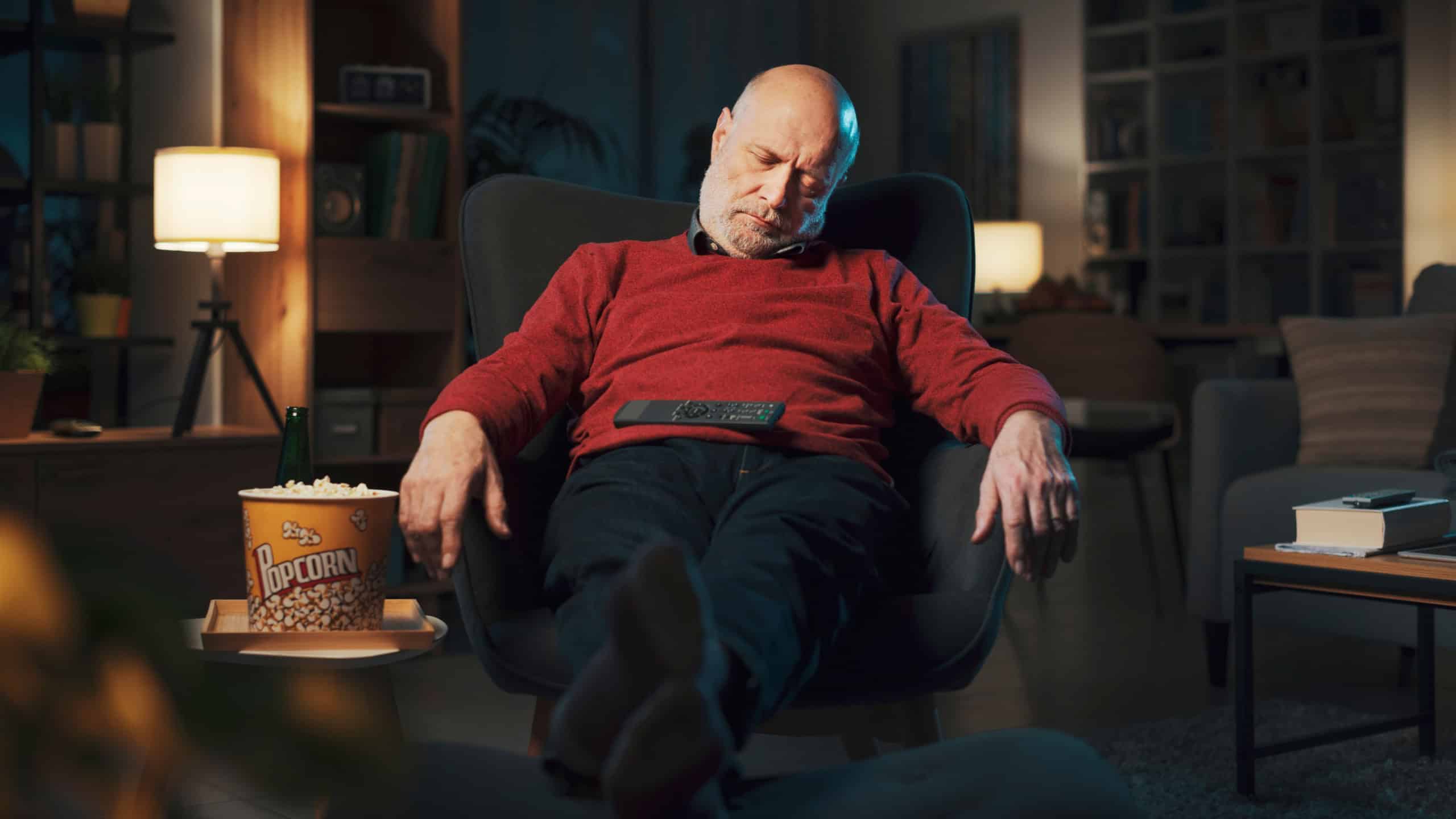 Tired senior man falling asleep in front of the TV late at night with the remote control on his belly