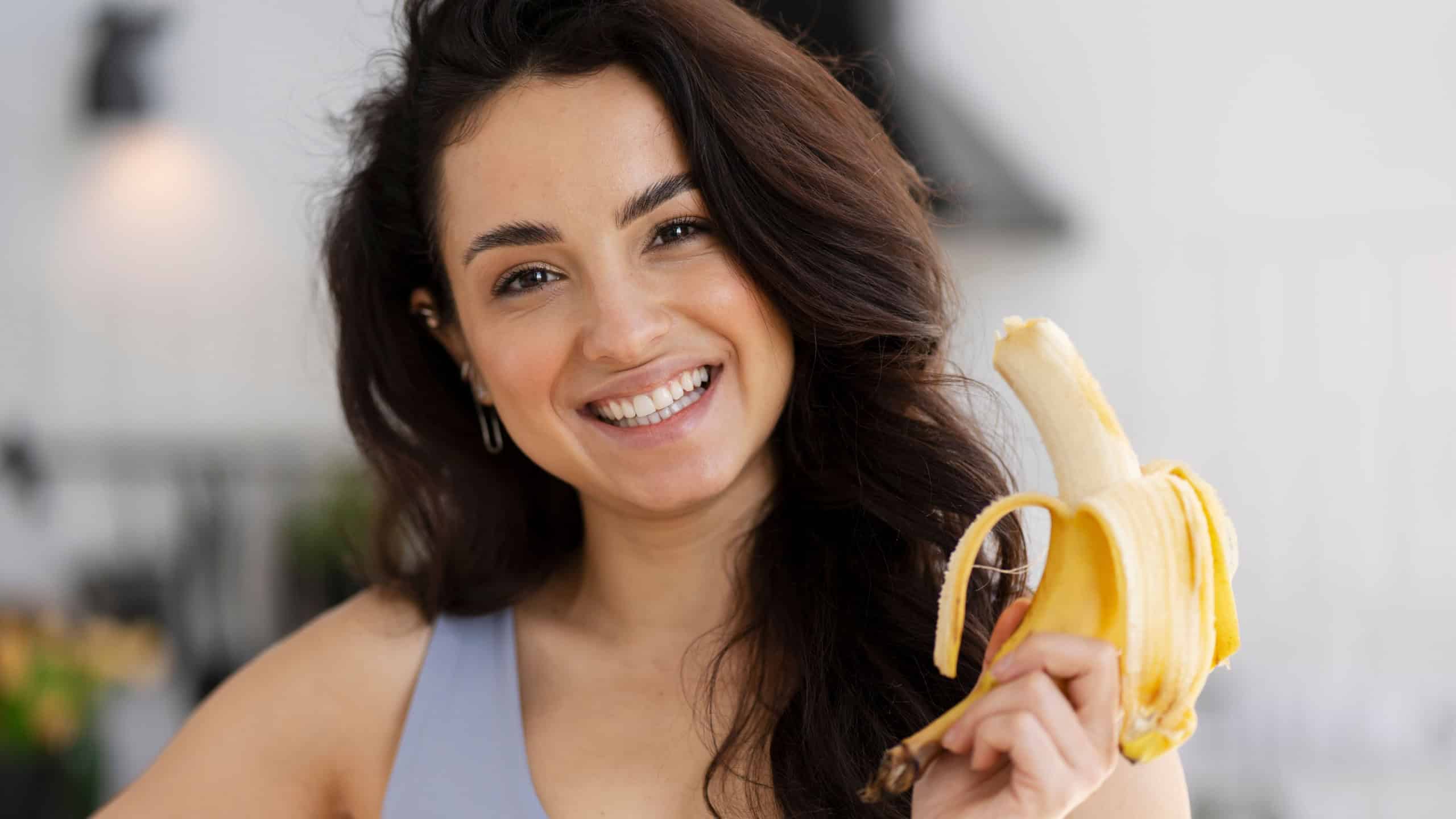 Portrait of diet smiling woman holding banana near face standing in modern kitchen. Healthy lifestyle and vegetarian food concept