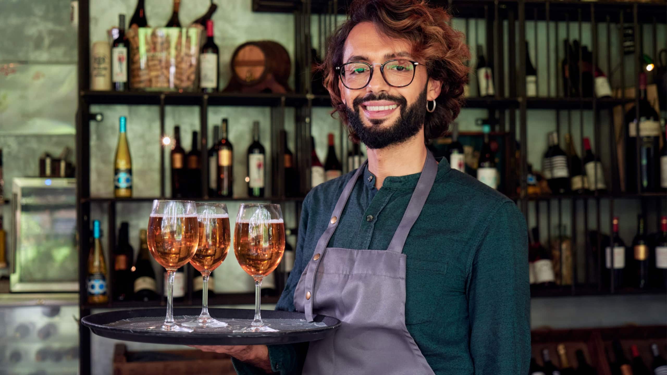 Portrait of a confident Italian waiter looking at camera while holding a tray with beers at his workplace. Entrepreneur young man at new restaurant. Copy space.
