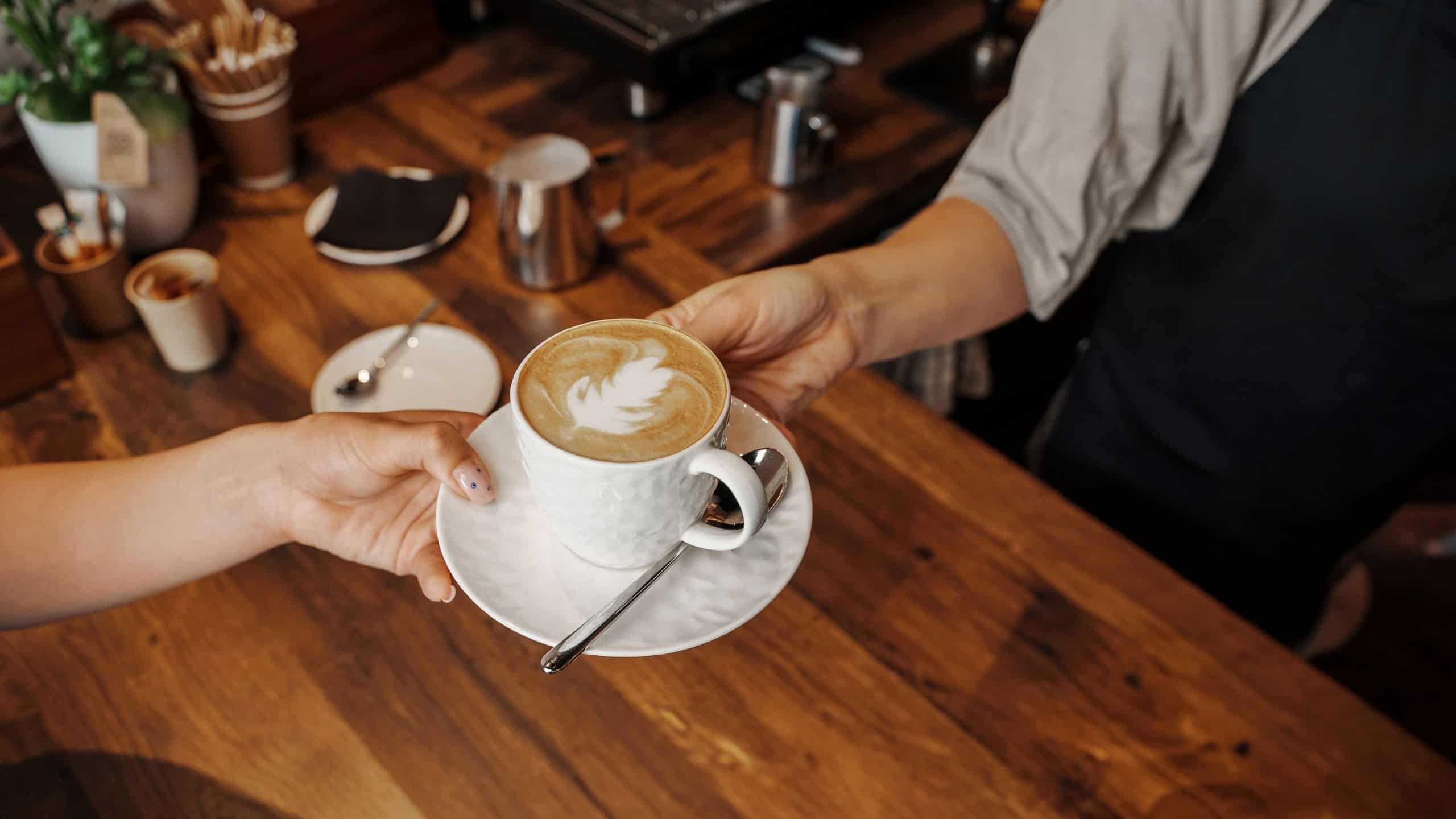 A barista hands over a beautifully crafted cappuccino with latte art to a customer at a wooden cafe counter