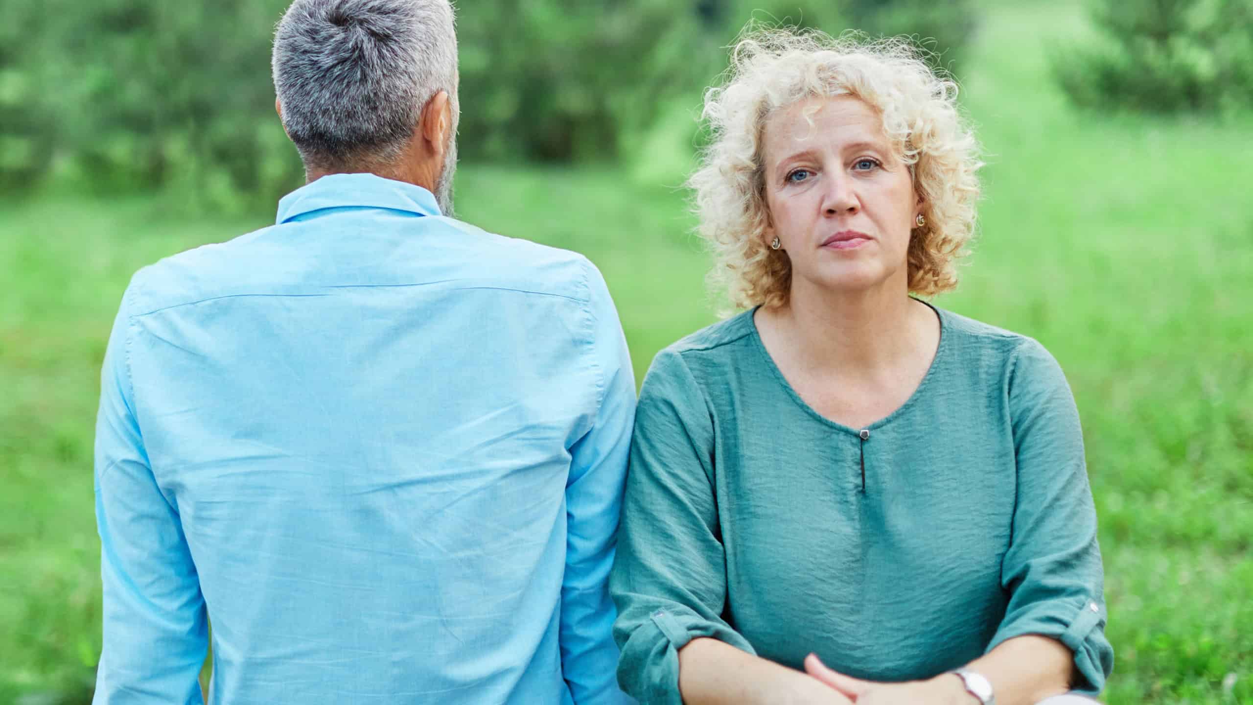 Mid age mature couple sitting on a park bench after a fight. Sad mature mid aged woman sitting with her back turned to her husband. Angry couple ignoring each other, relationship troubles