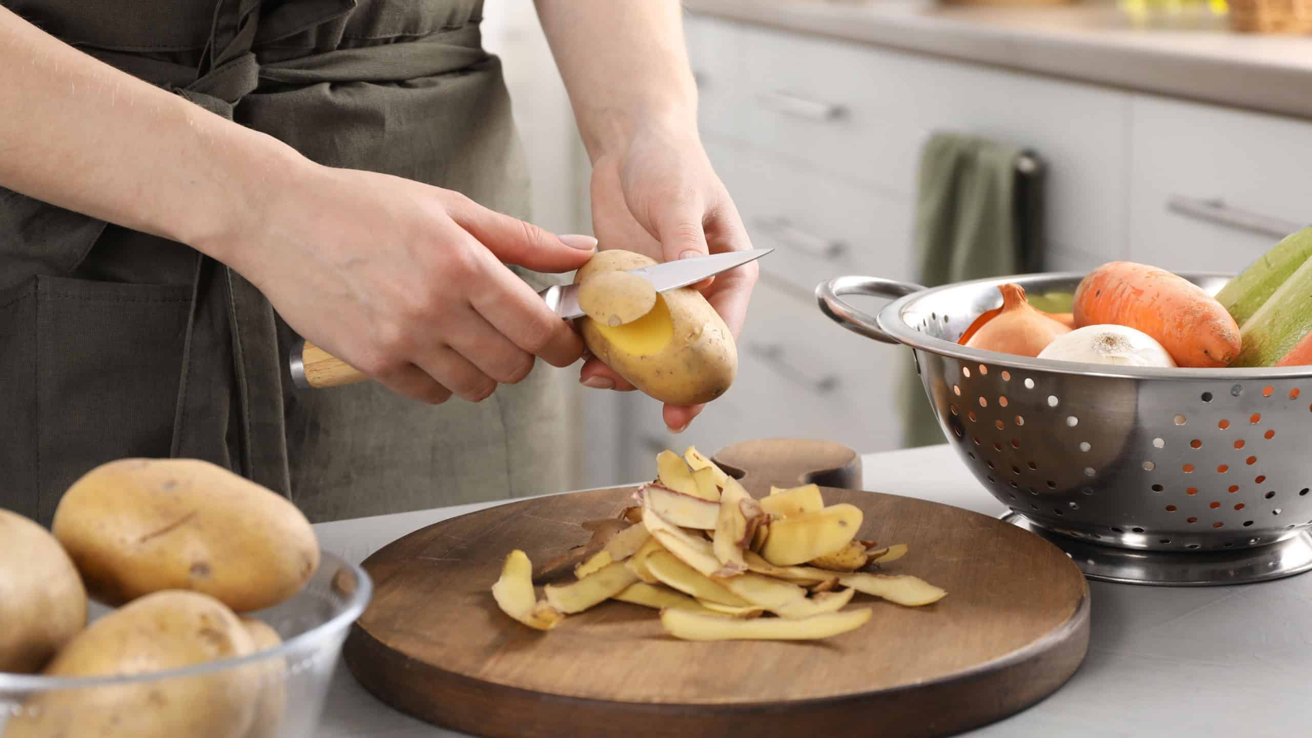 Woman peeling fresh potato with knife at light table indoors