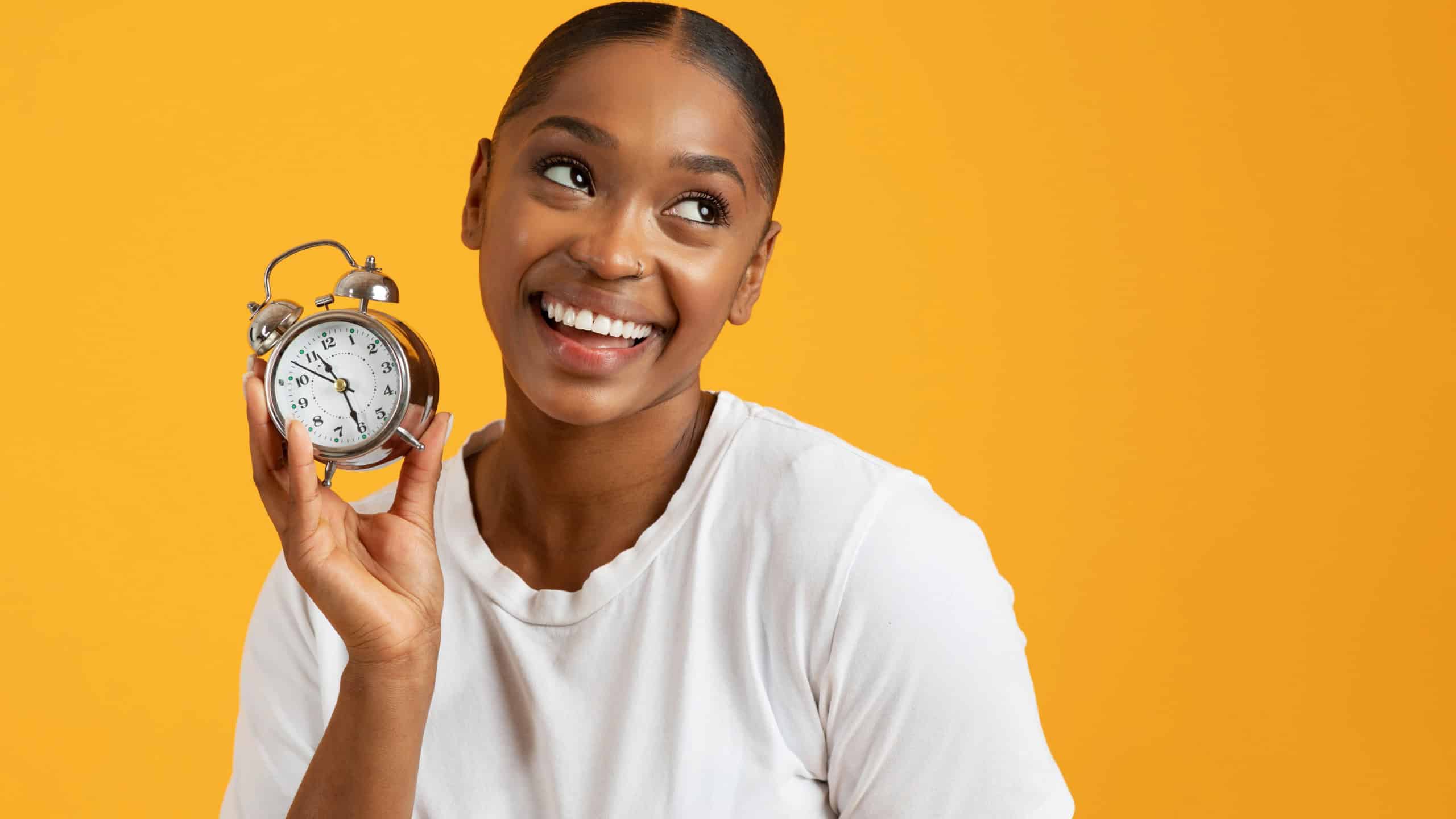 Cheerful black millennial woman holding alarm clock in hand with smile, reflecting concept of effective time management and punctuality, portrait on yellow background, free space
