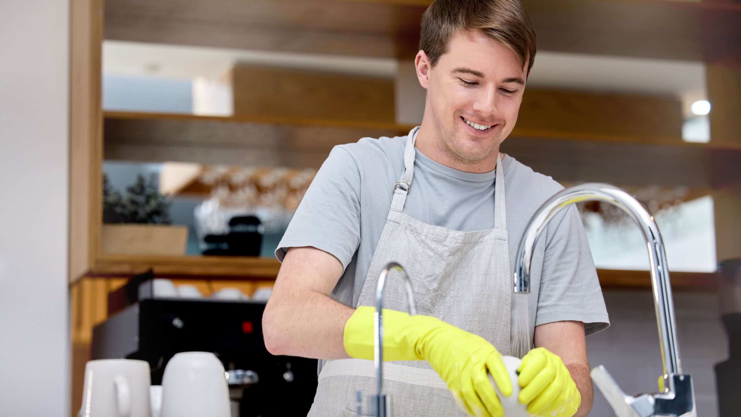 Cleaning, washing and dishes for man in home kitchen, chores and gloves for hand. Dirty, smile or hygiene with soap for bacteria prevention, germs and messy for disinfect crockery for male person