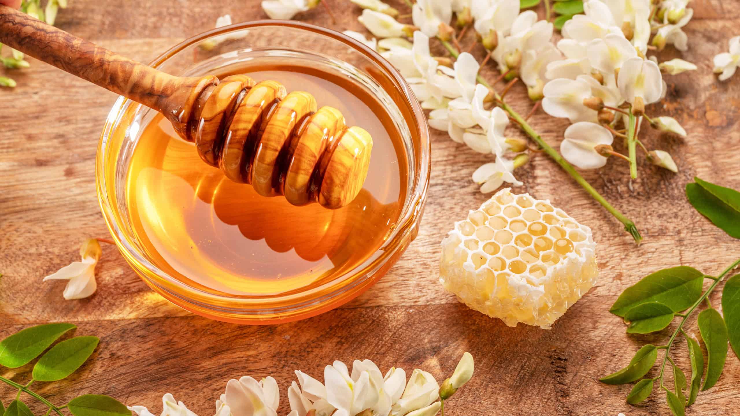 Honey dripping from wooden spoon in glass bowl with organic honey, honey plant acacia flowers and leaves near it on wooden table. Top view.