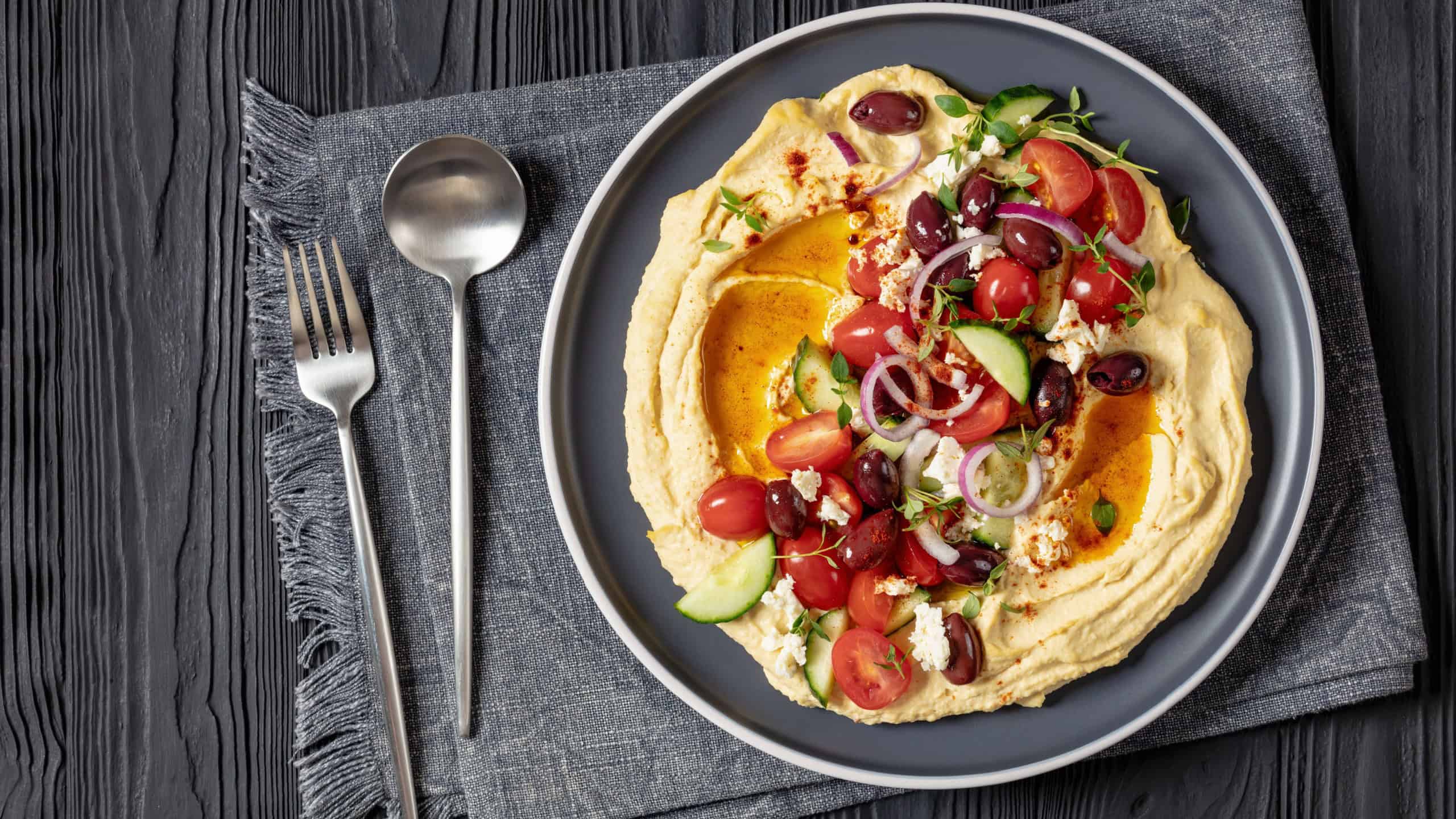 Loaded Greek hummus with fresh cucumbers, olives, tomatoes, feta cheese, red onions and thyme on plate on black wooden table with spoon and fork, flat lay