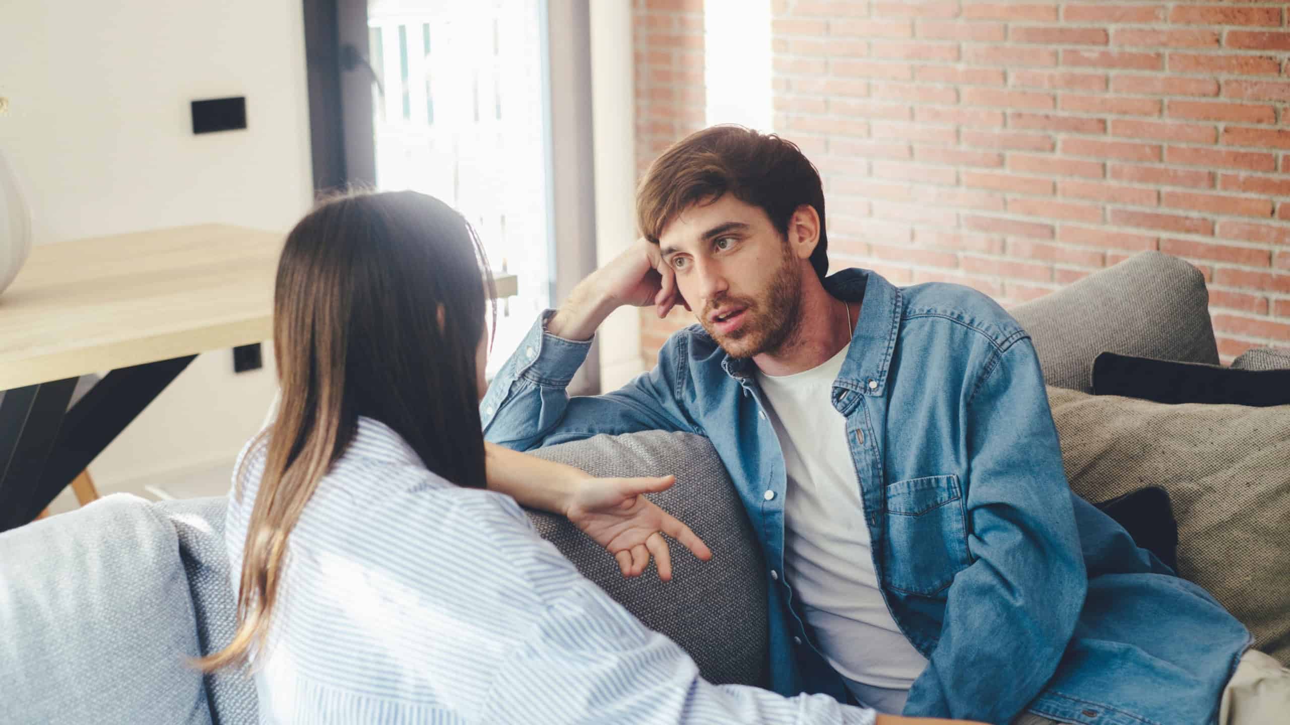 Serious young couple sitting together on sofa, talking about relationships, spending time together at home, concentrated husband listening to wife talking, friends having conversation