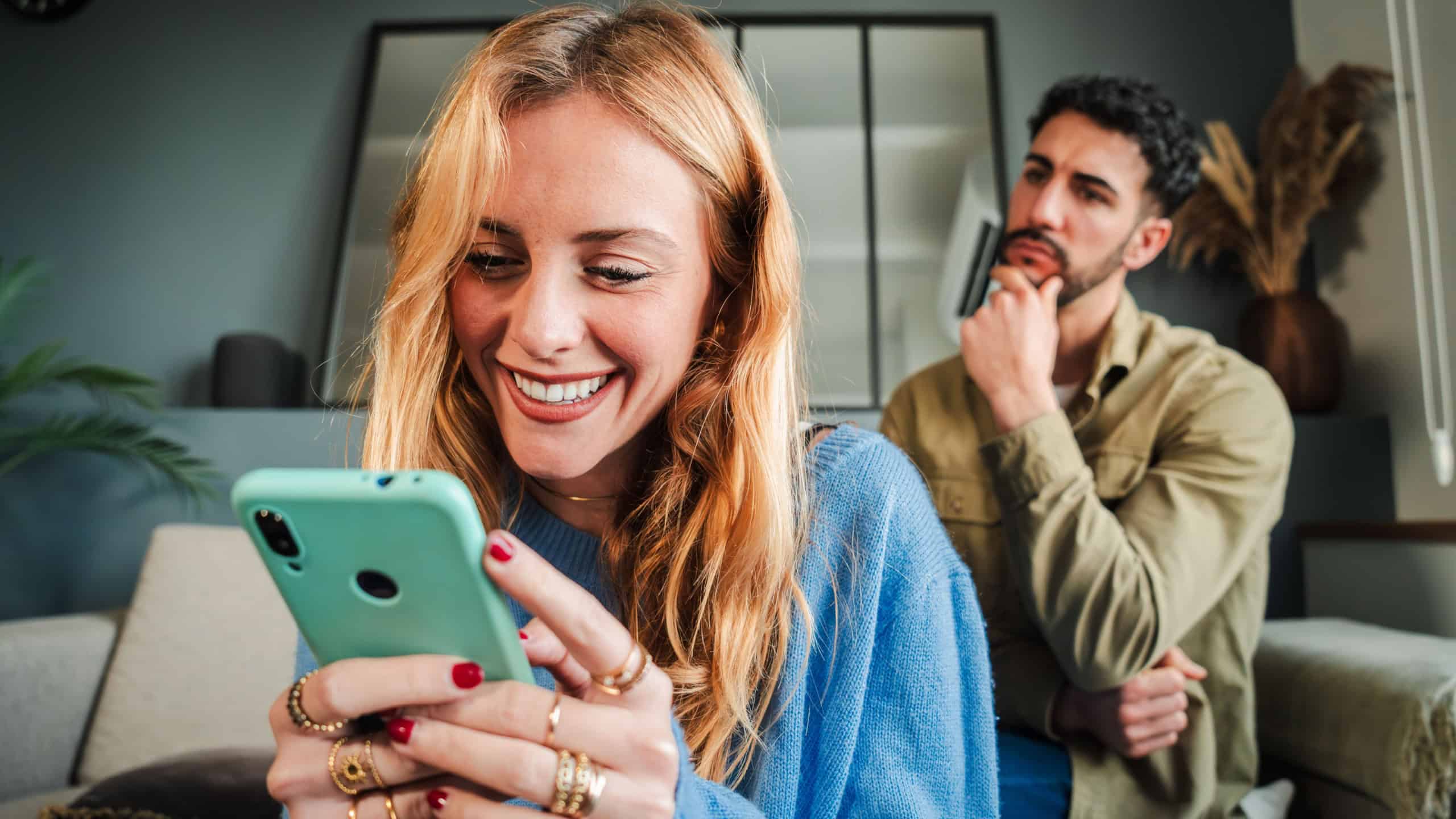 Woman secretly texting on her phone to deceive her husband, while he peeks over her shoulder and reads her messages. The boyfriend, feeling angry and upset, watches his girlfriend as she pays him no