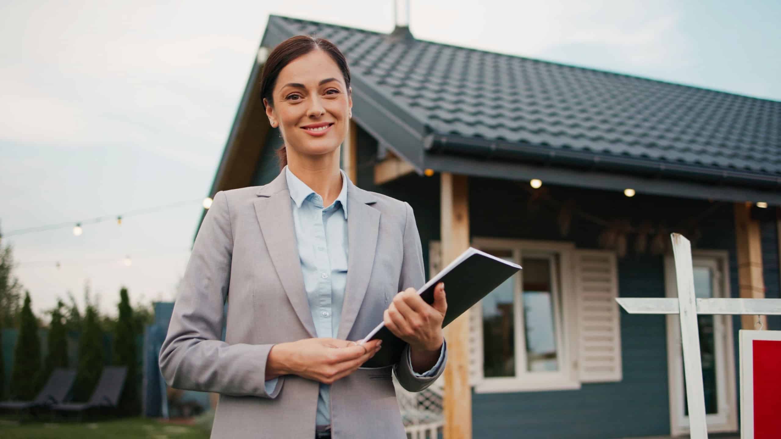 Beautiful Caucasian woman wearing official clothing. Making open arms gesture. Working as real estate agent. Cozy small house in background. Sign next to female showing property for sale.