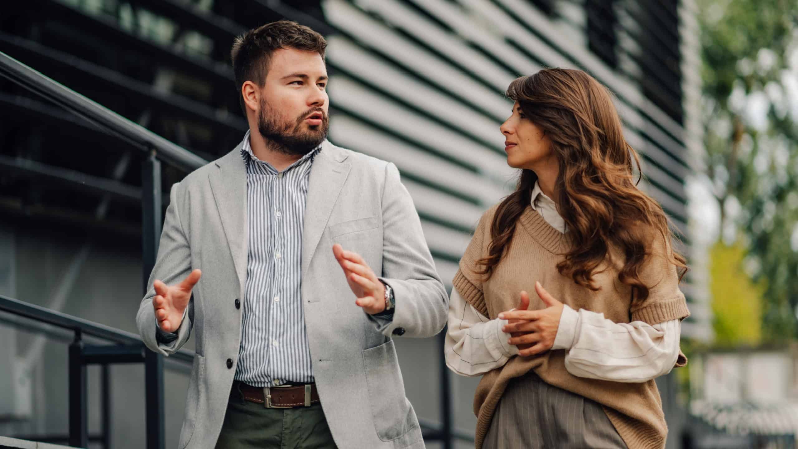 Two business partners having a conversation while walking outdoors in a business district, the man is gesturing while explaining something to his female colleague