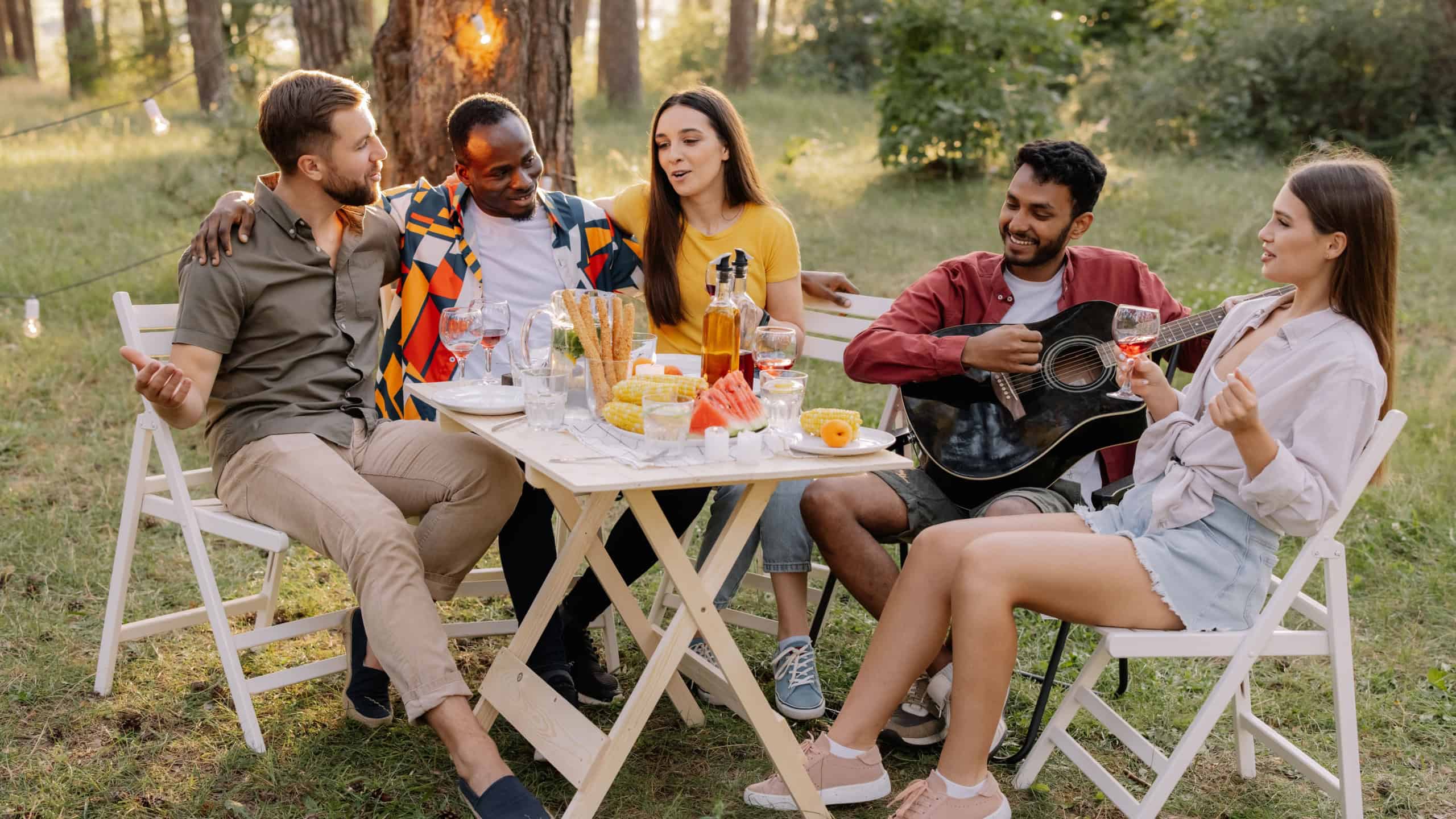 Meeting of multiracial group of friends playing guitar, singing, eating dinner and drinking wine during party in the forest
