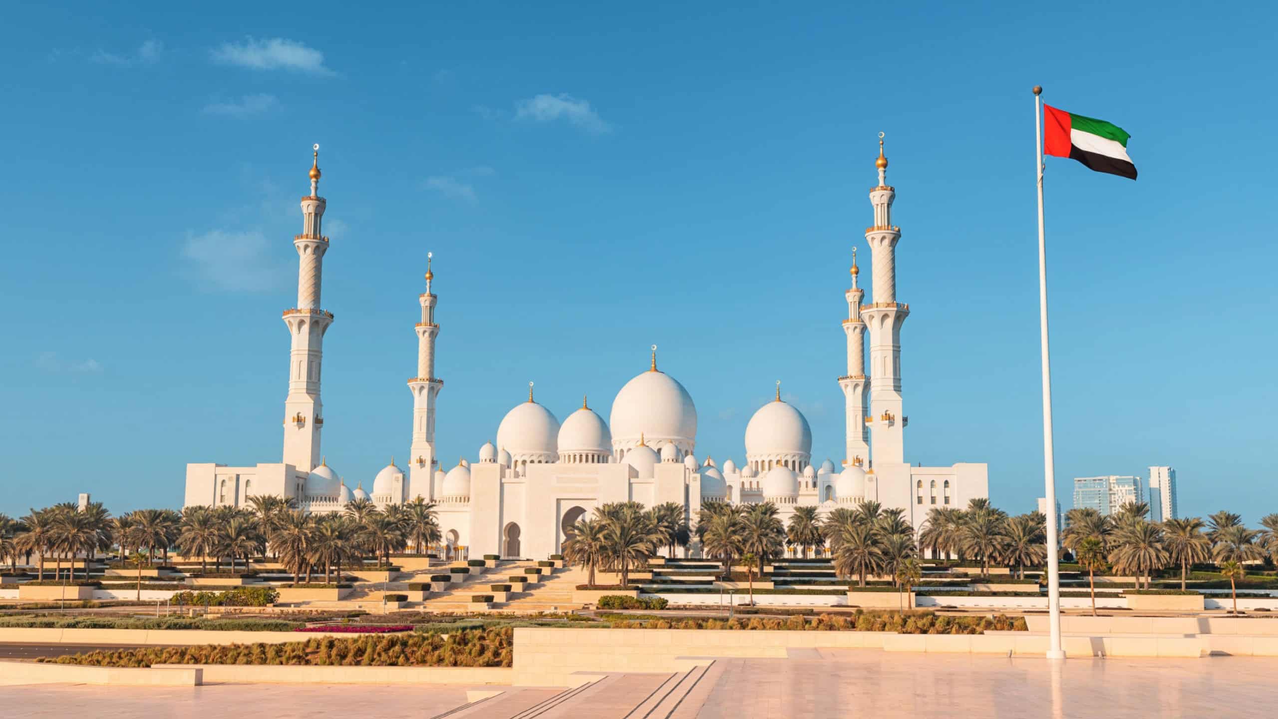 Scenic view of Sheikh Zayed Grand Mosque under a clear blue sky with the United Arab Emirates flag waving proudly