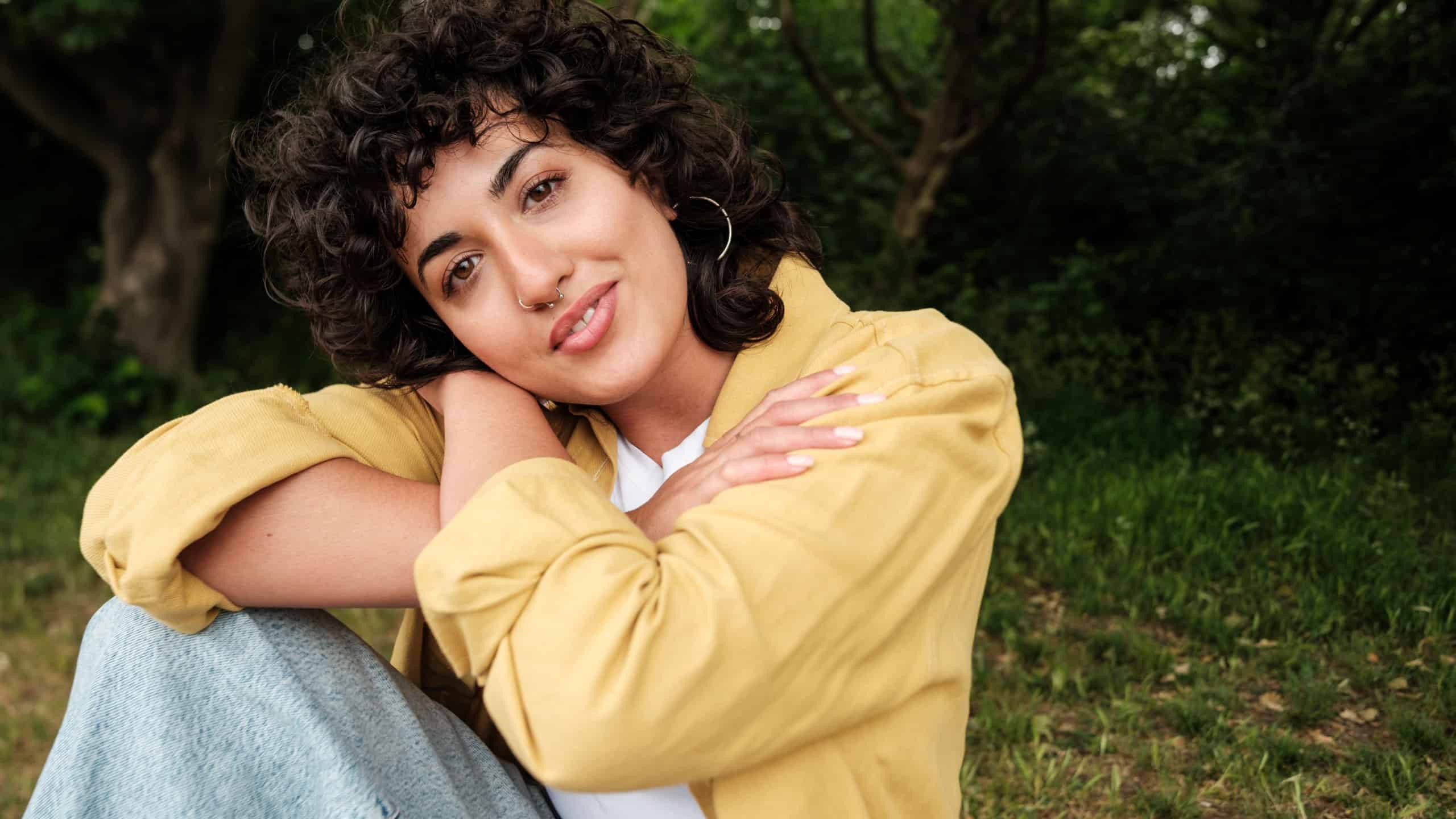 Sitting in a serene natural setting, a young woman embraces herself with a gentle smile, radiating confidence, self-acceptance, and a deep connection with nature