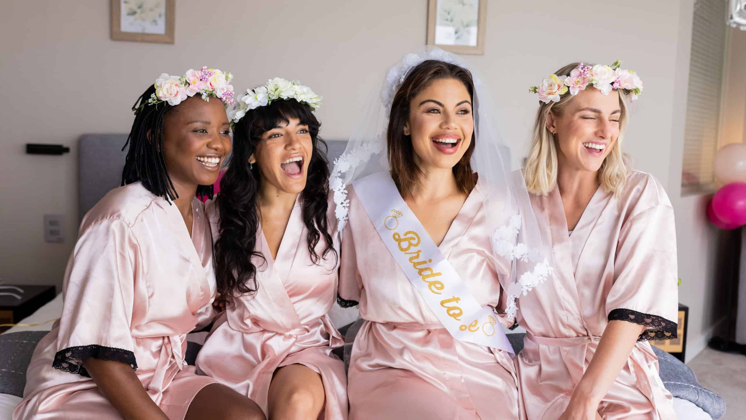 Bride-to-be with friends wearing robes and flower crowns, laughing on bed. Bachelorette, celebration, friendship, bridal party, floral, pajamas