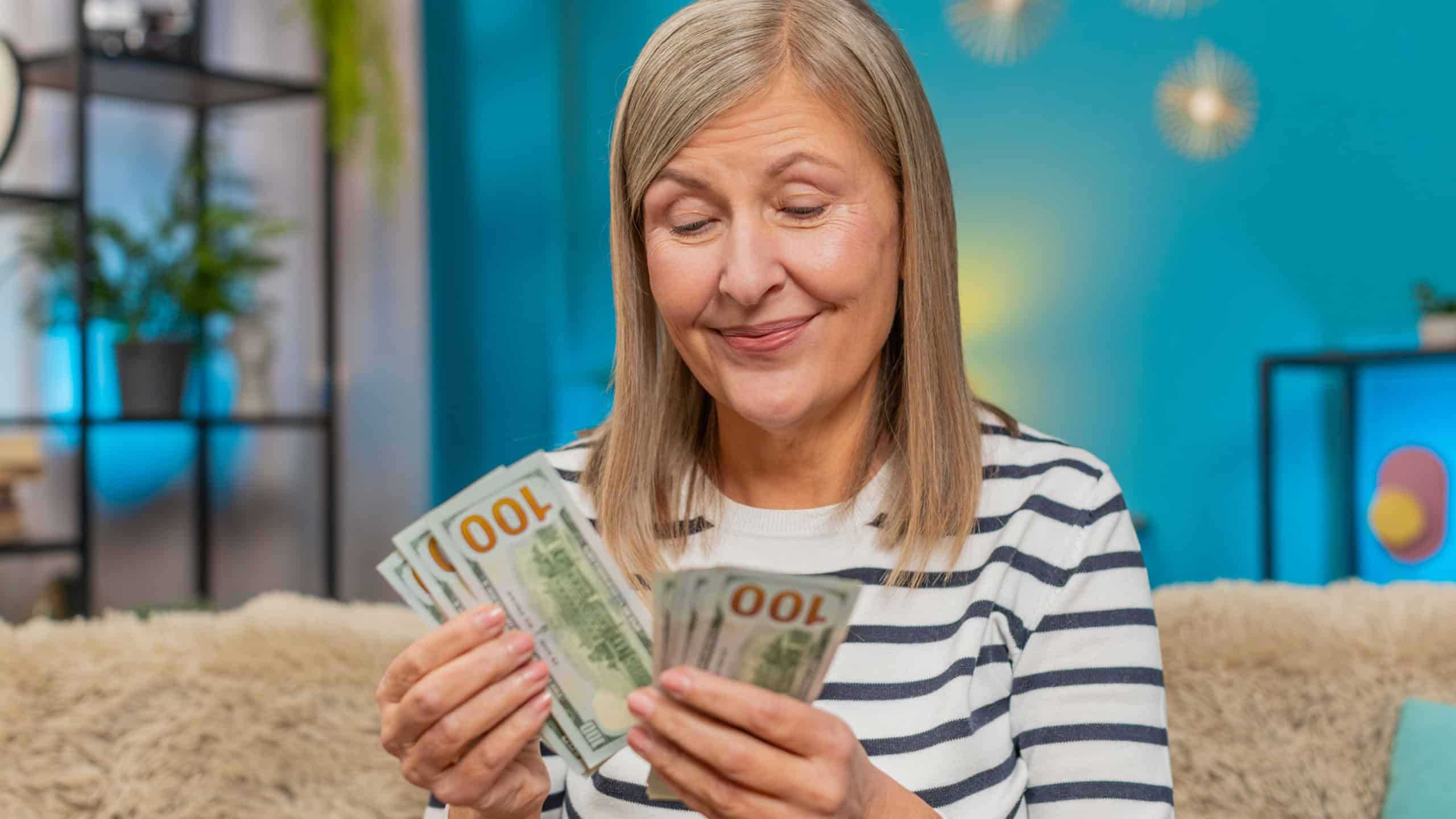 Happy Caucasian mature woman counts money dollar cash in hands after winning lottery bonus or receiving a gift. Grandmother smiles broadly sitting on home sofa, celebrating financial victory salary