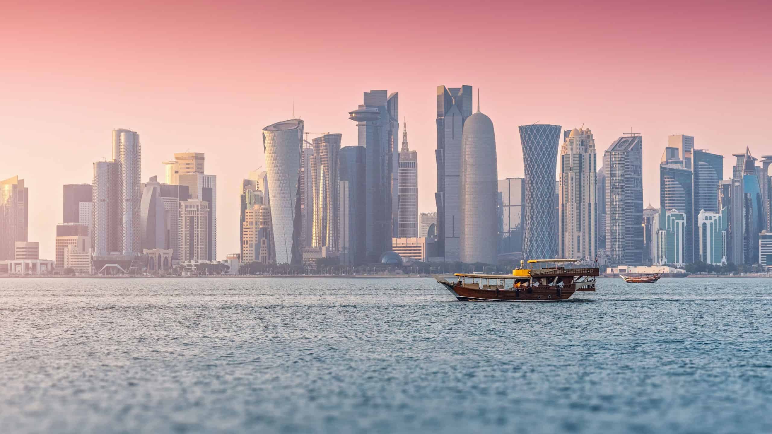 Doha city skyline featuring West Bay skyscrapers along the Persian Gulf during a beautiful pastel sunset, with dhow boats sailing
