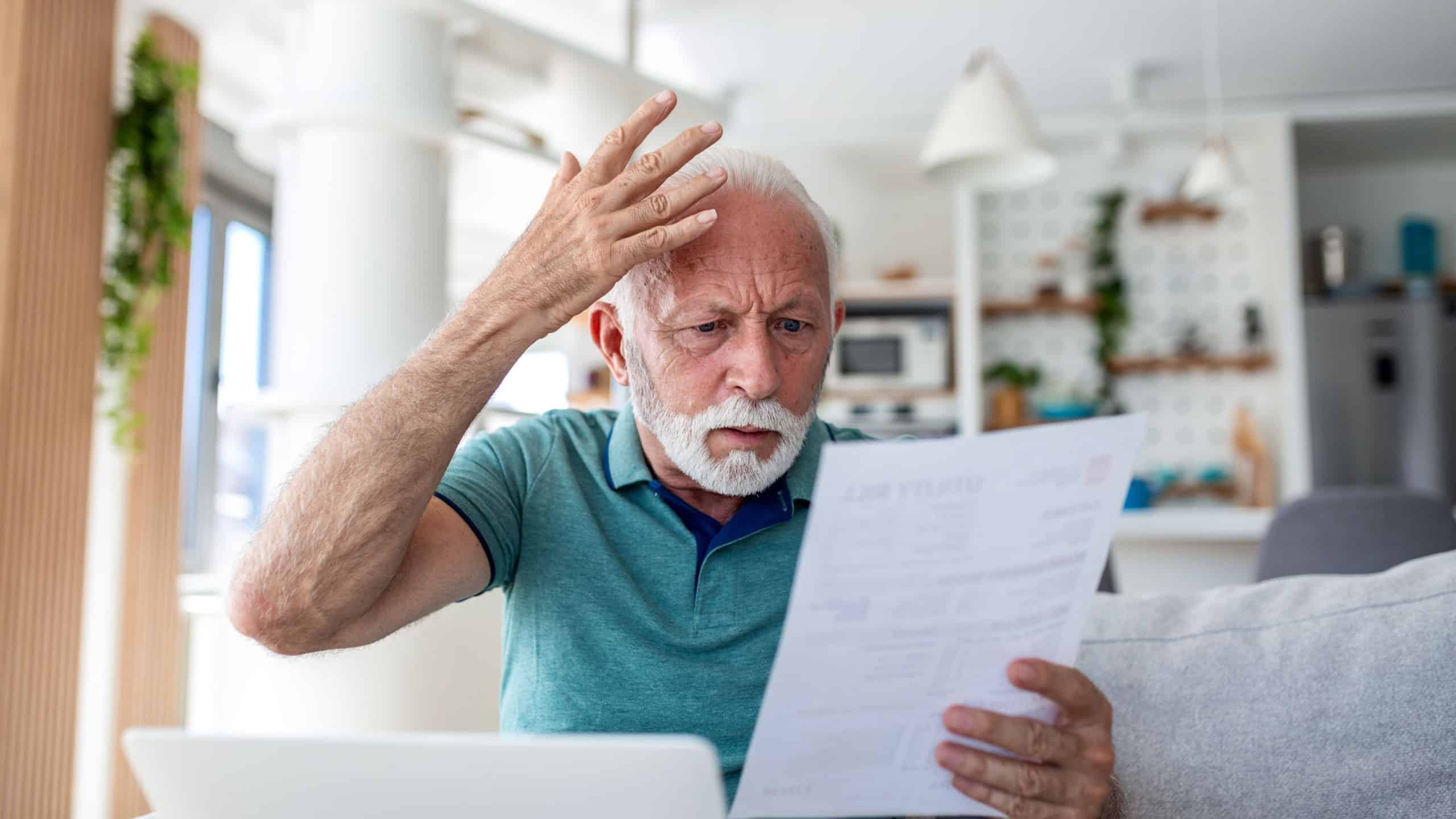 Worried senior man reading utility bill and using laptop at home. Stressed elderly male looking at paper with debt or high taxes. Financial problem, bankruptcy and money crisis concept for seniors.