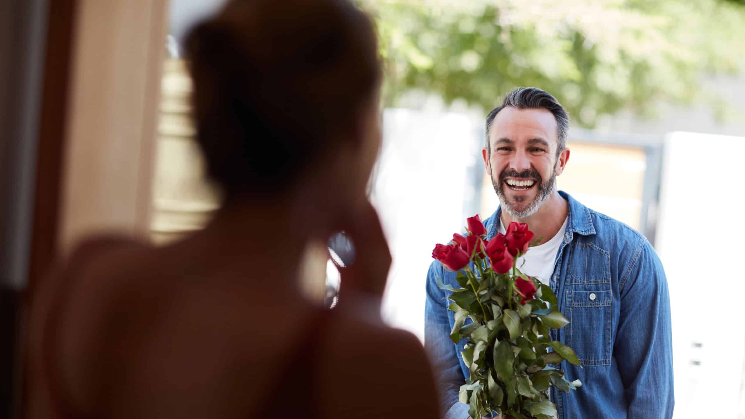 Roses, couple happy man with surprise by front door, valentines day gift and anniversary celebration. Flower bouquet, love and people with romantic present for marriage, home and partner support