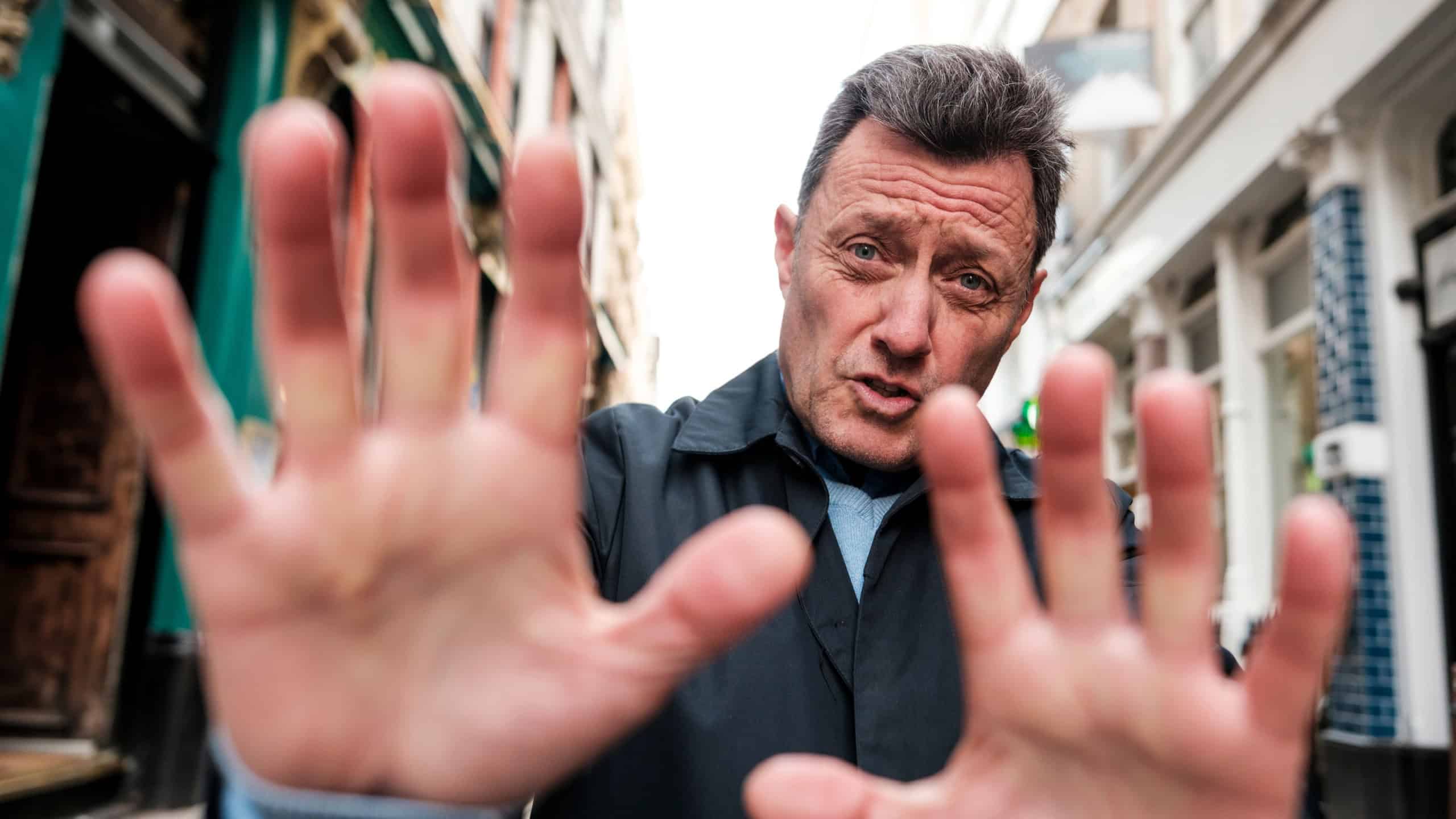 Mature man standing in a narrow city street, facing camera with open palms raised in a stop gesture, expressing refusal, boundary and desire for personal space