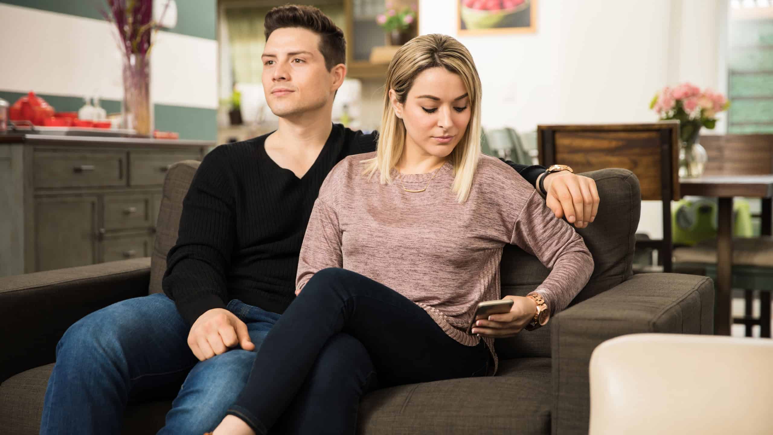 Pretty young woman sending a text to her lover while sitting next to her boyfriend at home