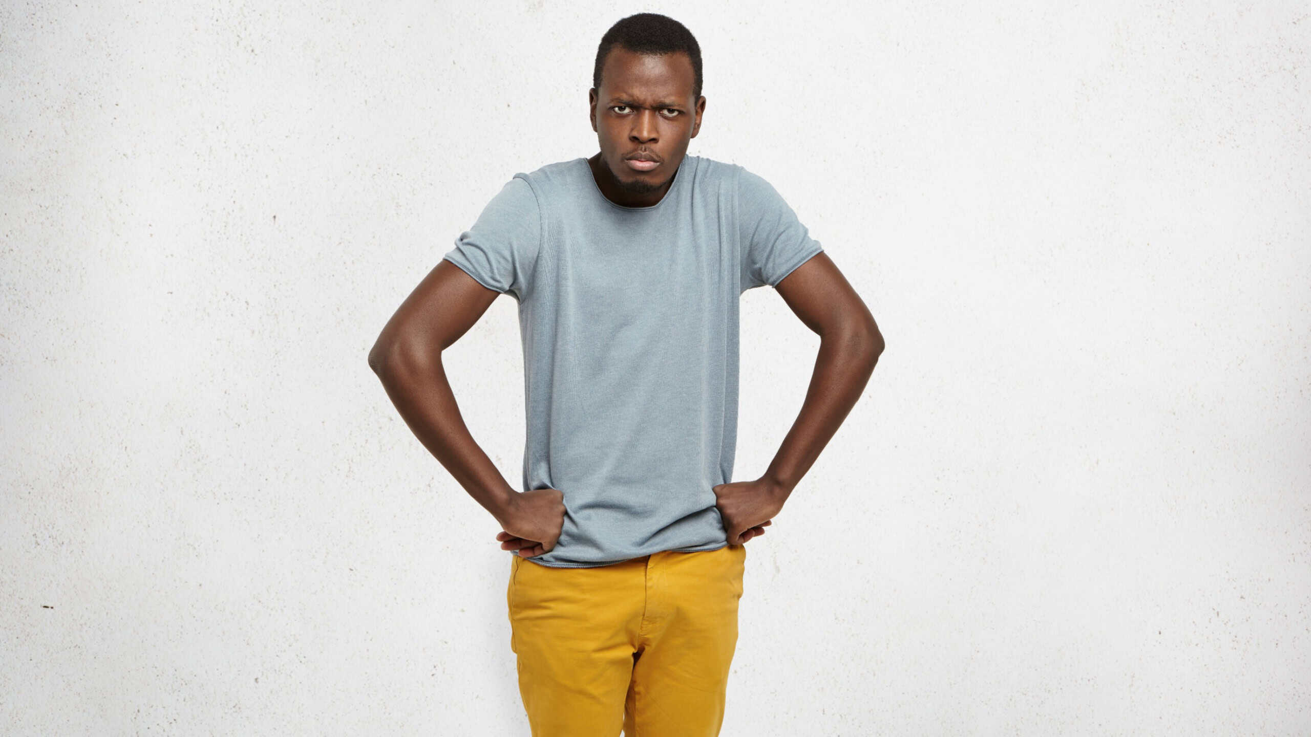 Cropped portrait of mad African American male wearing grey t-shirt and mustard jeans holding hands on hip, frowning, having grumpy angry look, dissatisfied with misbehavior of his little children