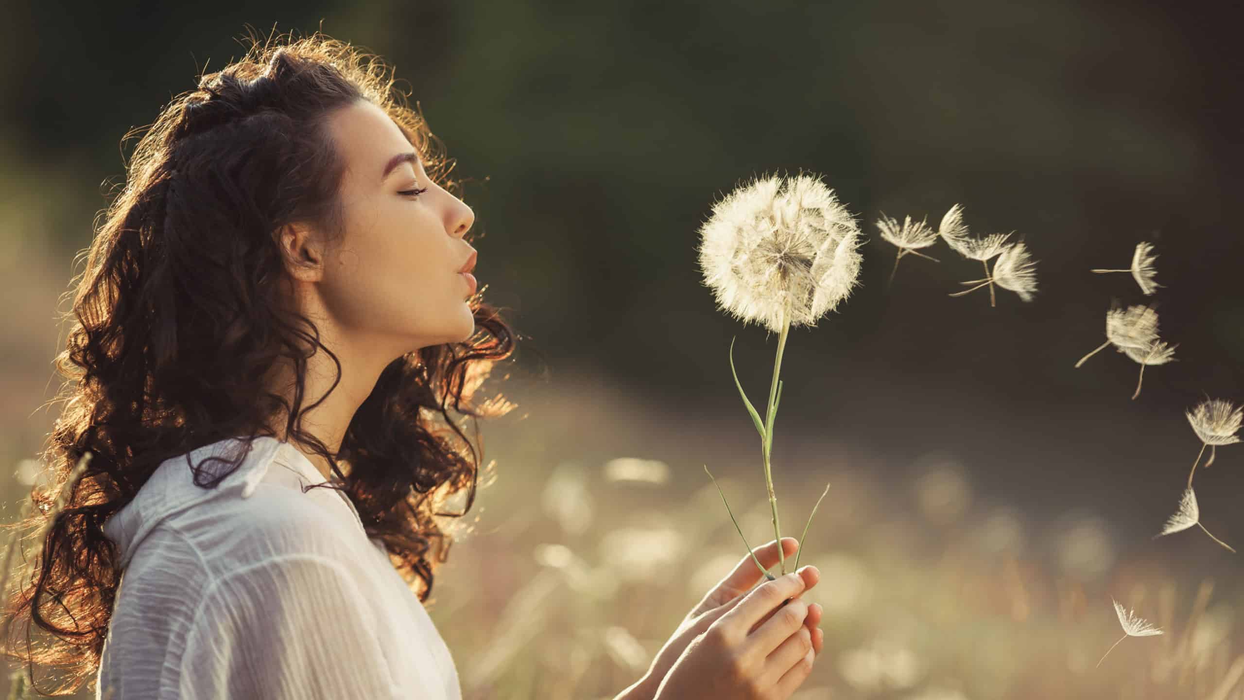 Beautiful Young Woman sitting on the field in green grass and blowing dandelion. Outdoors. Enjoy Nature. Healthy Smiling Girl on summer lawn. Allergy free concept. Gorgeous slim mixed race Caucasian