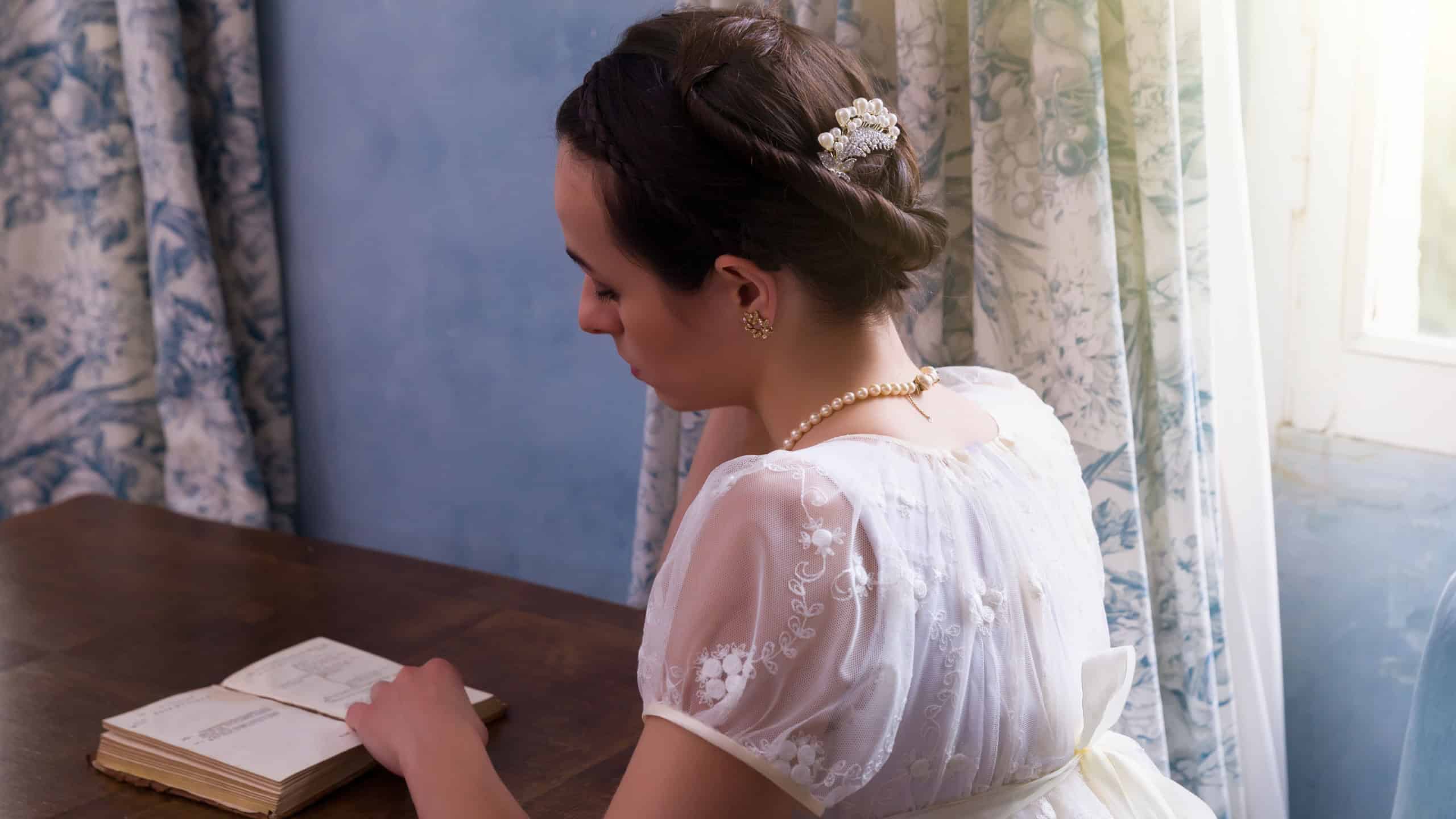 Pretty young woman in authentic regency dress reading a book in vintage room