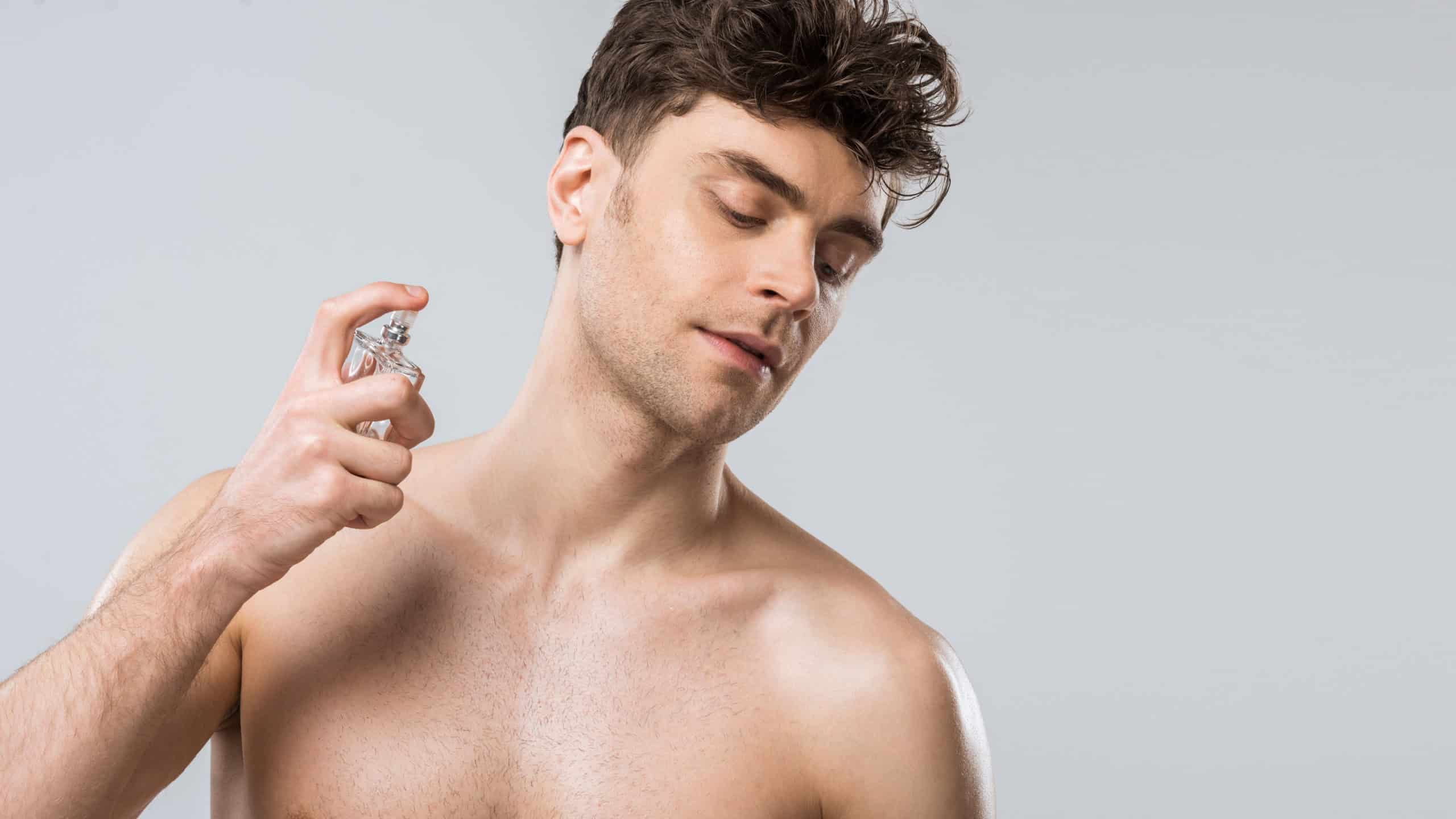 Shirtless sexy young man spraying perfumes, isolated on grey